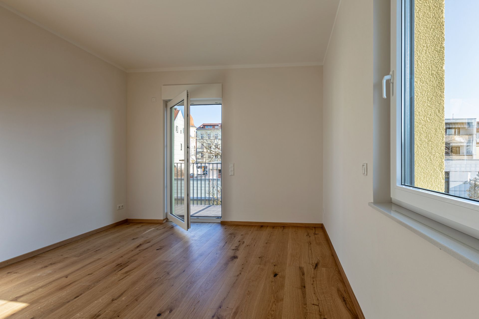 Leerer Raum mit Holzfußboden, offenem Fenster und Zugang zu einem kleinen Balkon.