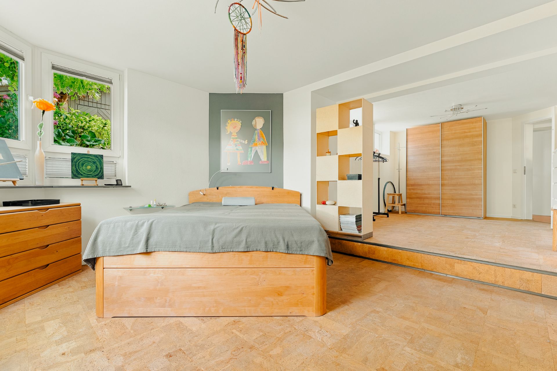 Helles Schlafzimmer mit Holzbett, Kommode, Regal und großen Fenstern, Parkettboden.