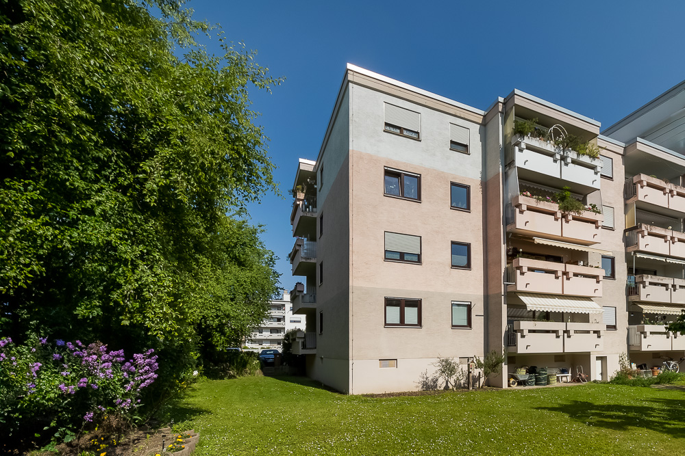 Wohnhaus mit mehreren Balkonen, Grünfläche und Bäumen im Vordergrund.