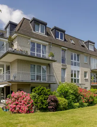 Modernes Mehrfamilienhaus mit Balkonen, umgeben von grünem Garten und blühenden Büschen.