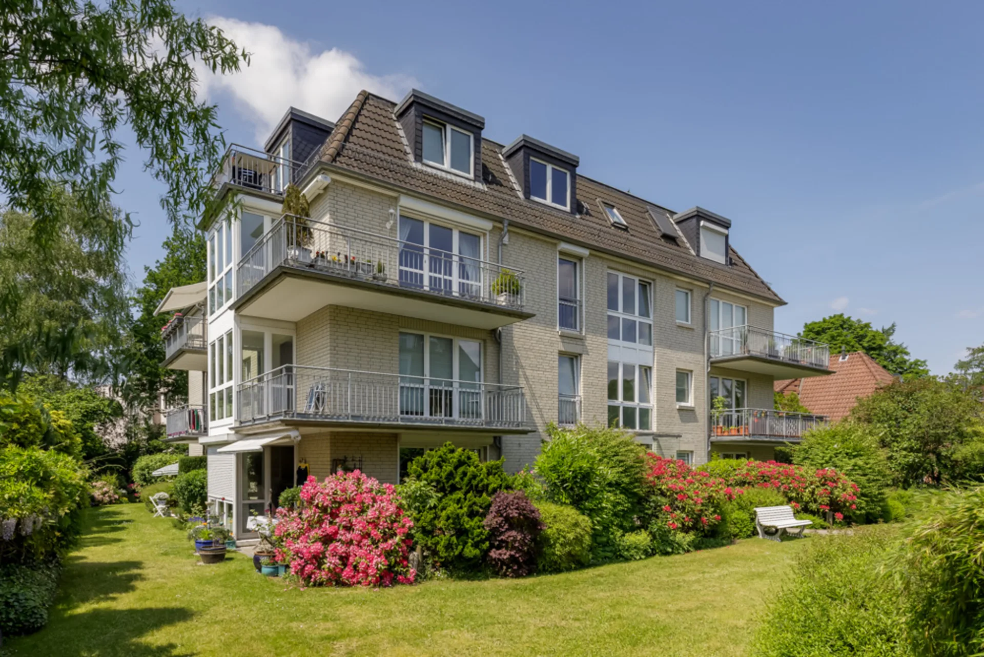 Modernes Mehrfamilienhaus mit Balkonen, umgeben von grünem Garten und blühenden Büschen.