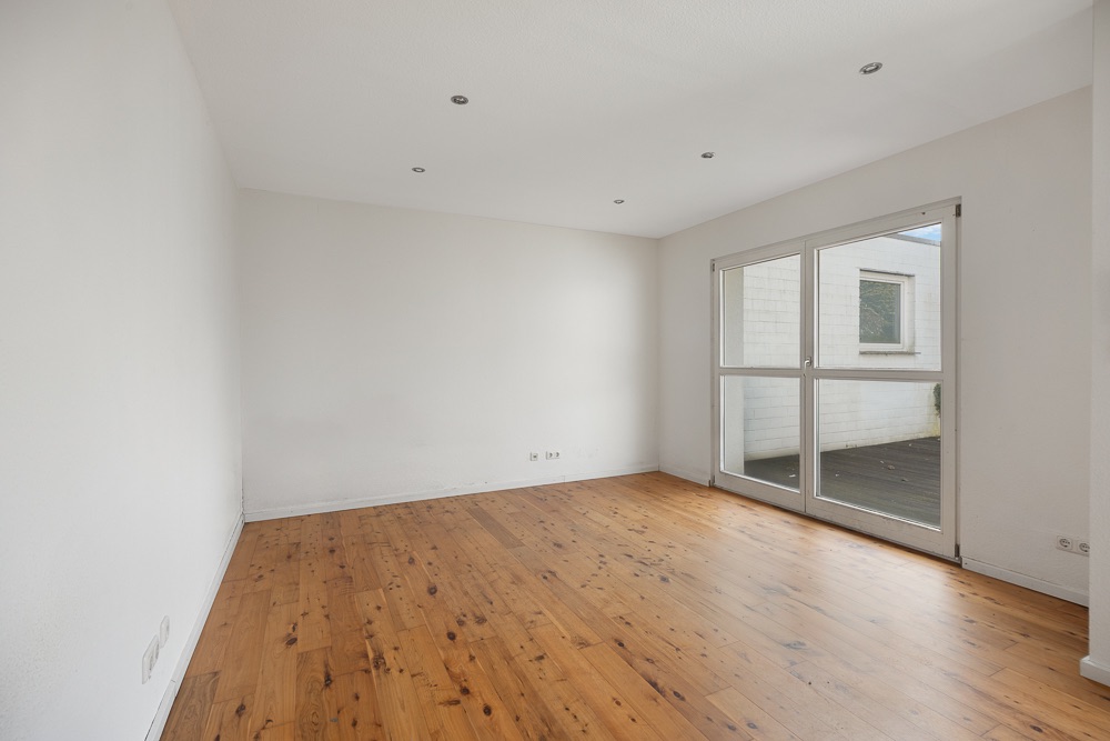 Leerer Raum mit Holzboden, weißen Wänden und großen Fenstern zur Terrasse.