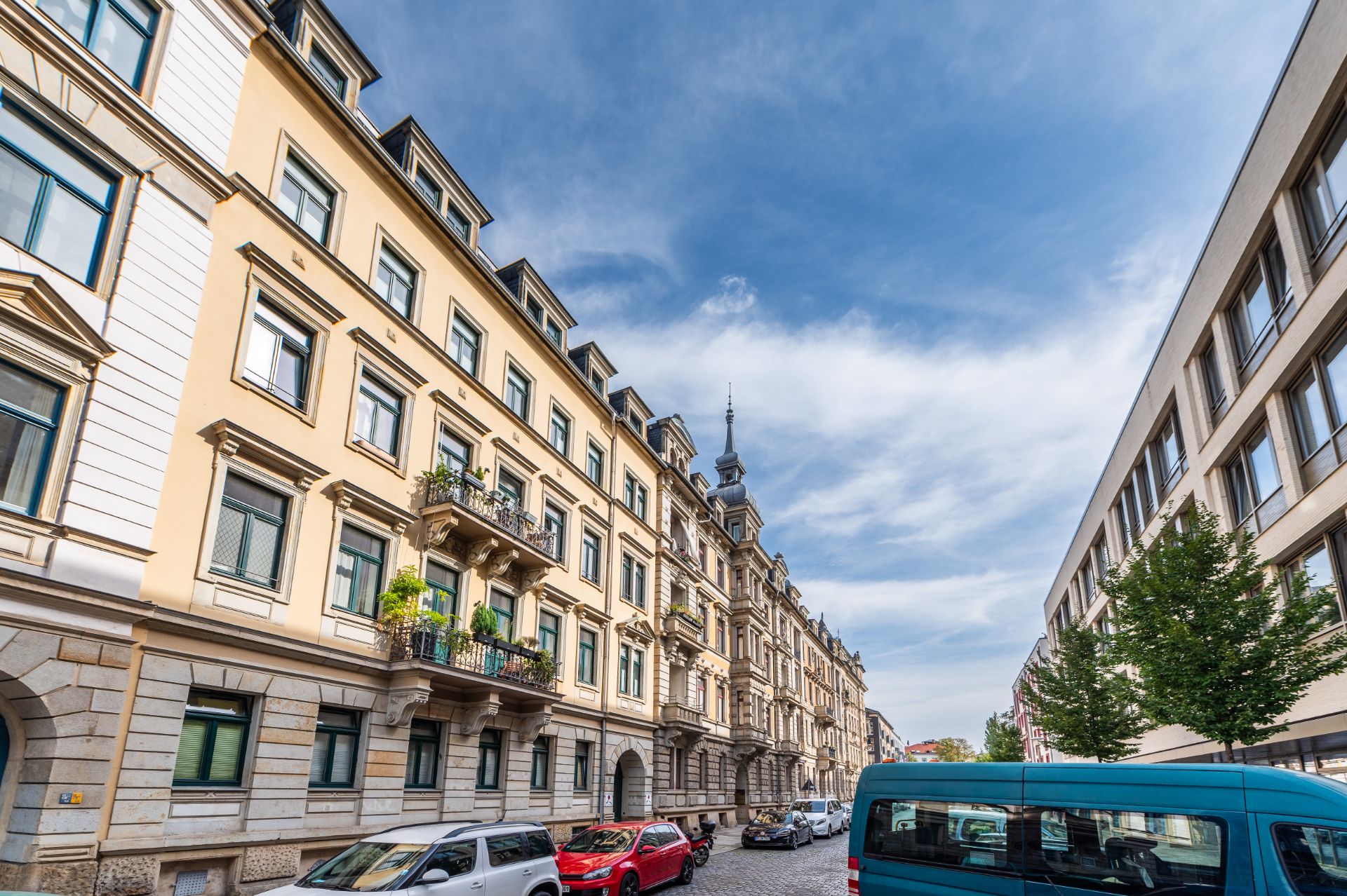 Wohngebäude mit Balkonen an einer Straße, geparkte Autos und ein blauer Himmel.