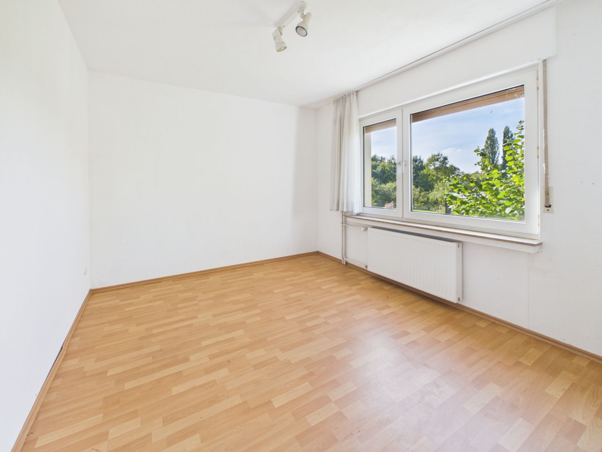 Leerer Raum mit Parkettboden, weißen Wänden und großem Fenster mit Blick ins Grüne.