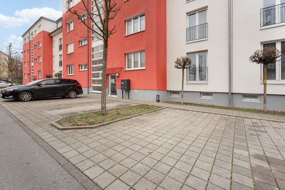 Eingang eines Wohngebäudes mit roter Fassade, Auto und gepflastertem Gehweg.