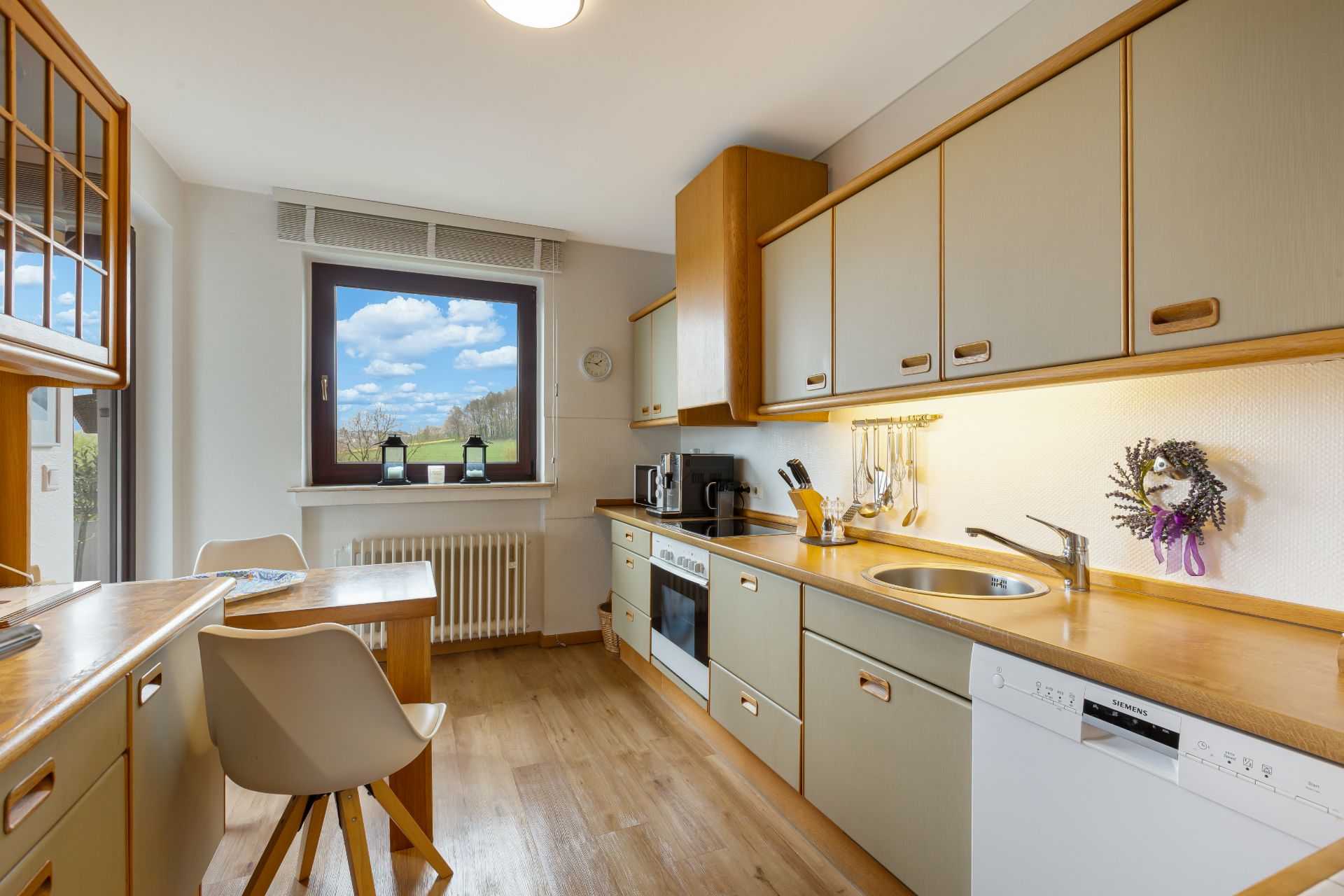 Helle Küche mit Holzfronten, Fenster, Esstisch, Stühlen und Küchenutensilien an der Wand.