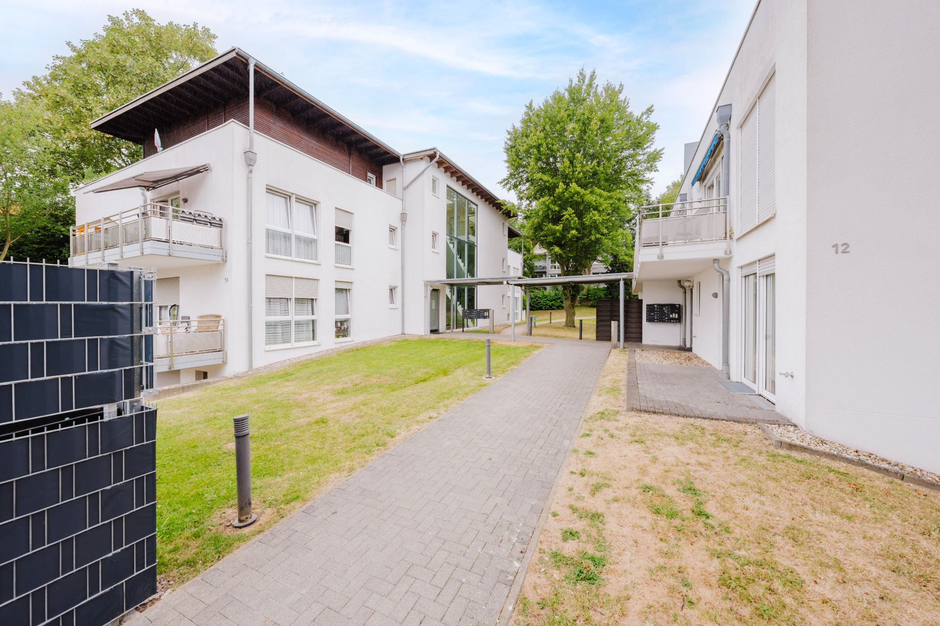Moderner Wohnkomplex mit weißen Gebäuden, Balkonen und Gehweg, umgeben von Bäumen und Rasen.