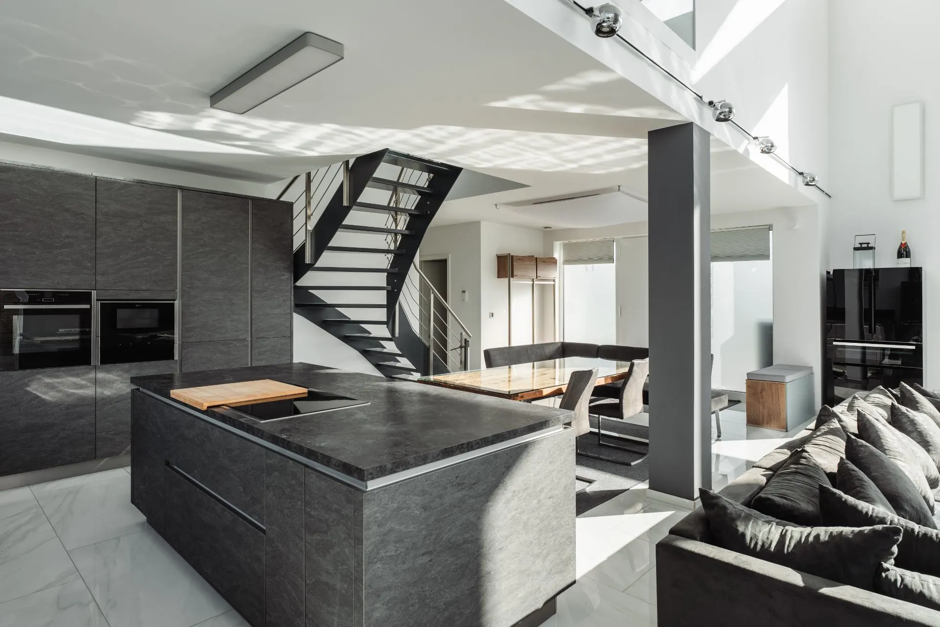 Moderne offene Küche mit dunkler Kochinsel, Esstisch, Sofa und Treppe im Hintergrund.
