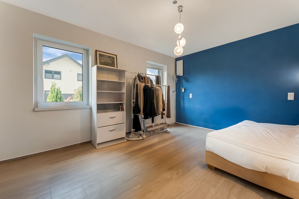 Helles Schlafzimmer mit blau gestrichener Wand, Bett, Regal, Kleiderständer und zwei Fenstern.