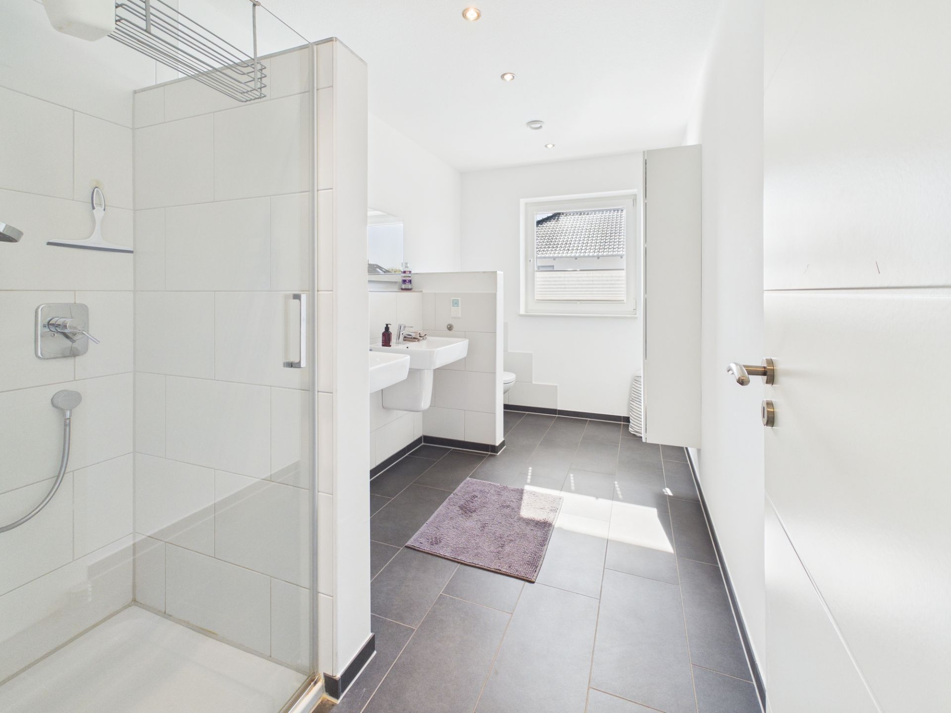 Helles Badezimmer mit Dusche, zwei Waschbecken und Fenster, dunkler Boden und weiße Wände.