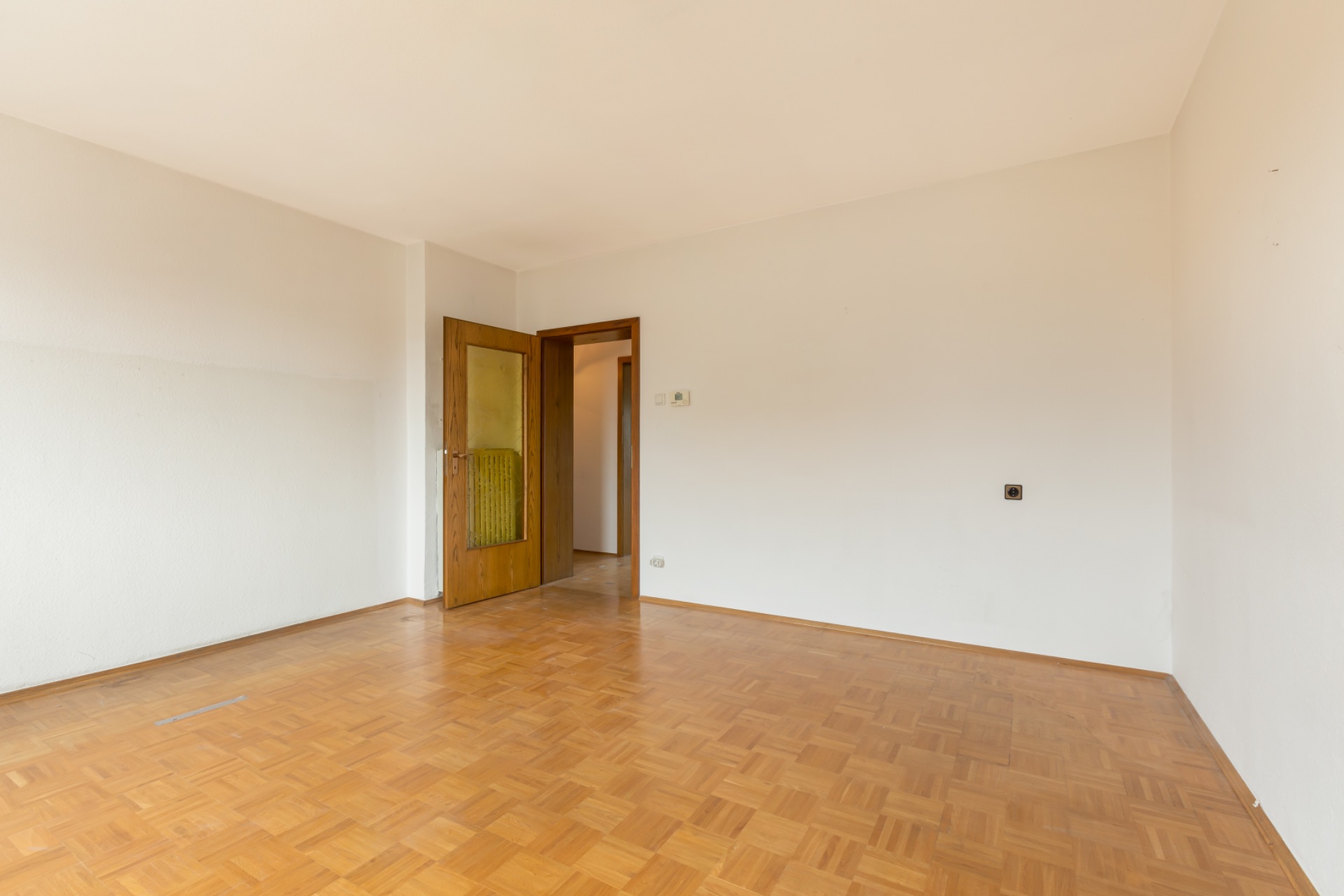 Leerer Raum mit Parkettboden, weißen Wänden und geöffneter Holztür.