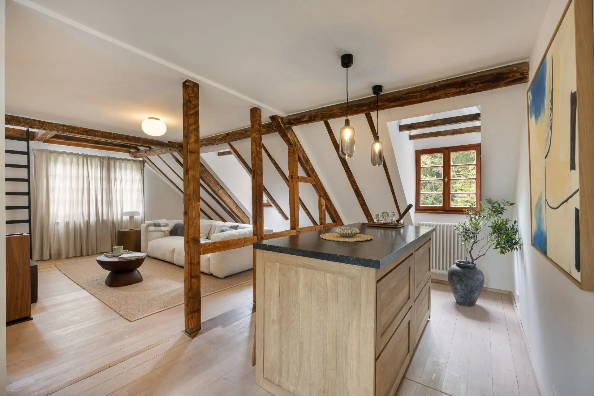 Helles Dachgeschoss mit Holzbalken, Küche, Wohnzimmer, großem Fenster und Pflanze im Topf.