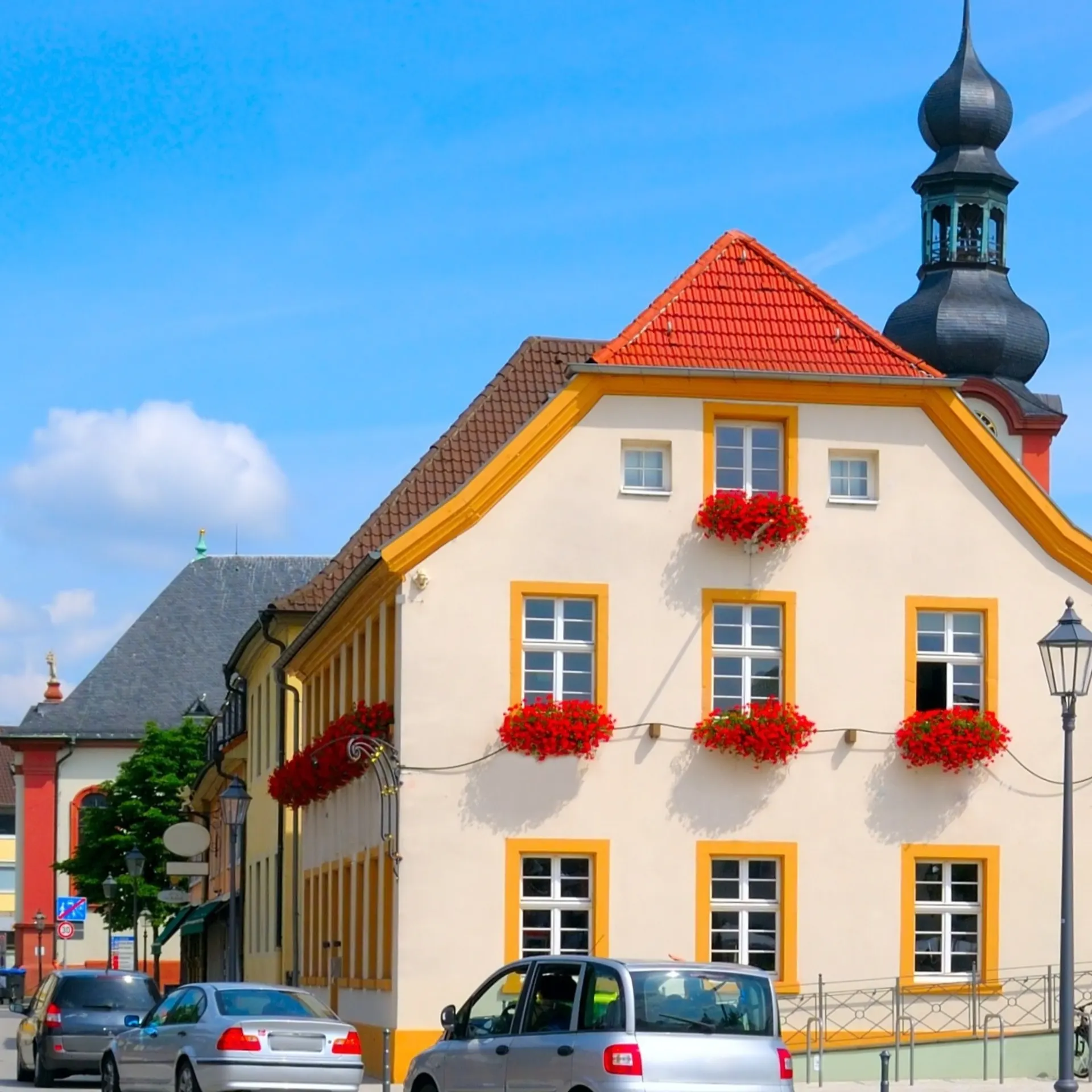 Wohnhaus mit roten Blumen an den Fenstern, Autos parken davor, blauer Himmel.