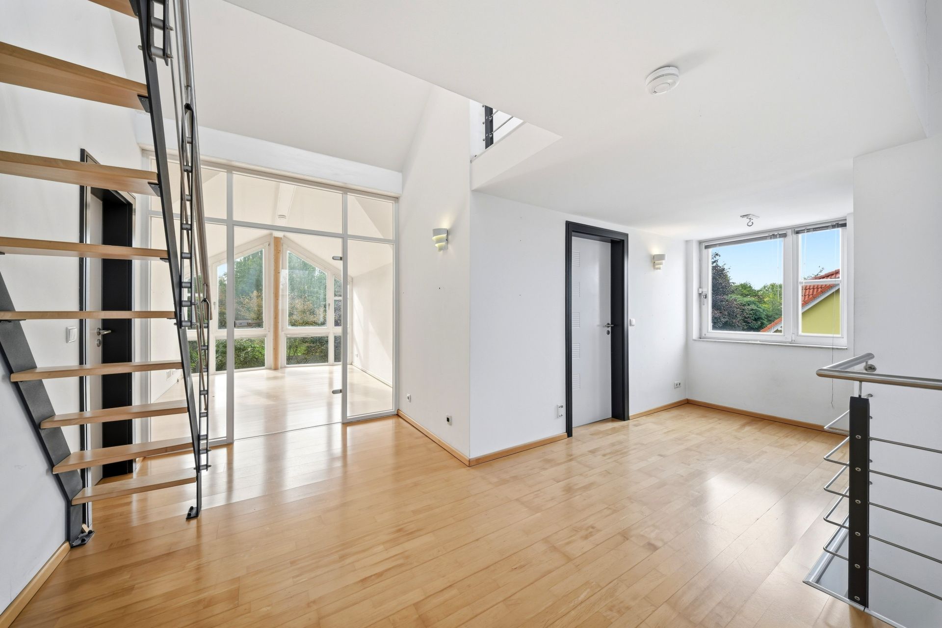 Helles Zimmer mit Holzfußboden, Treppe, großem Fenster und Glastüren zu einem weiteren Raum.