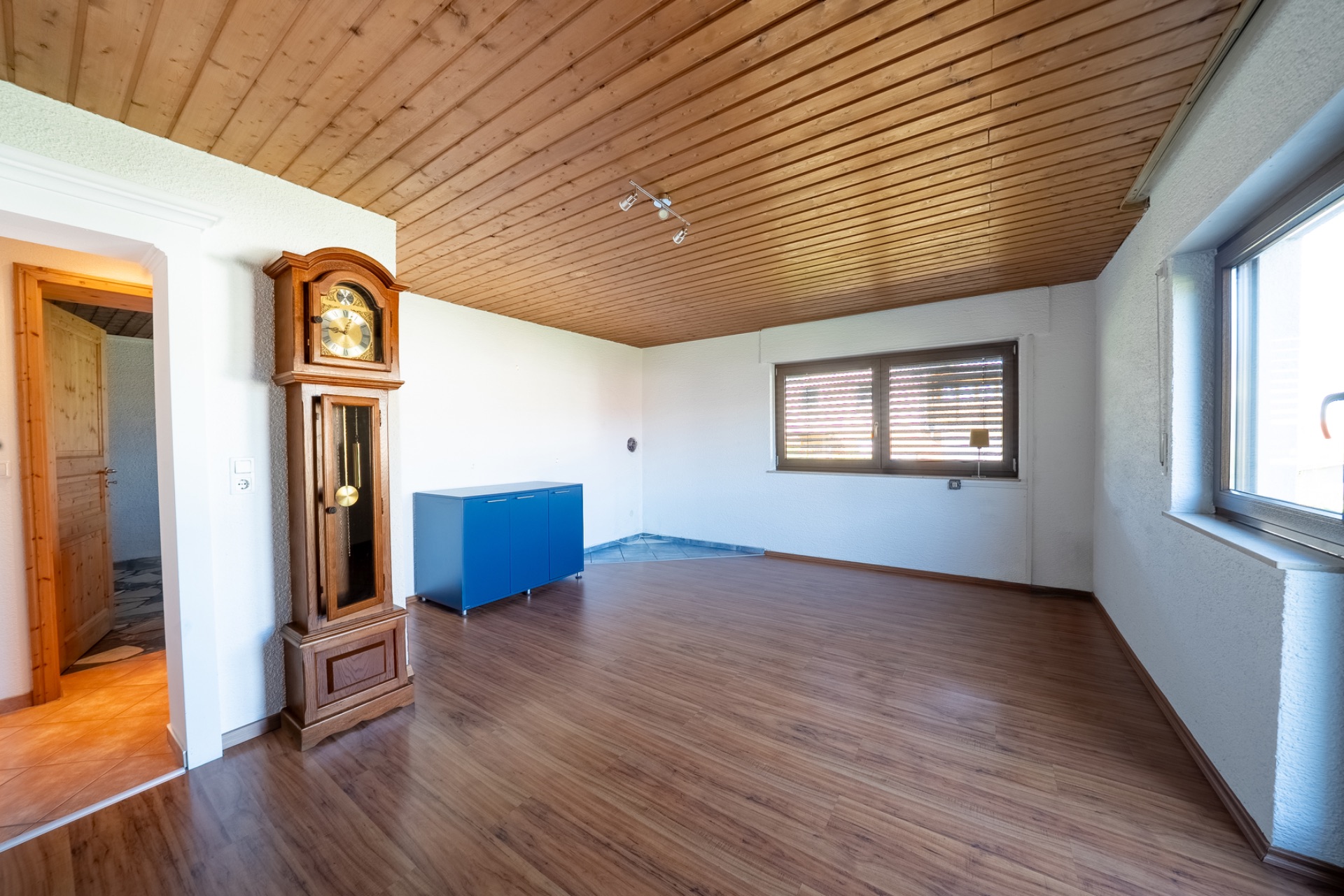 Leeres Wohnzimmer mit Holzdecke, Großvateruhr, blauem Schrank und großen Fenstern.