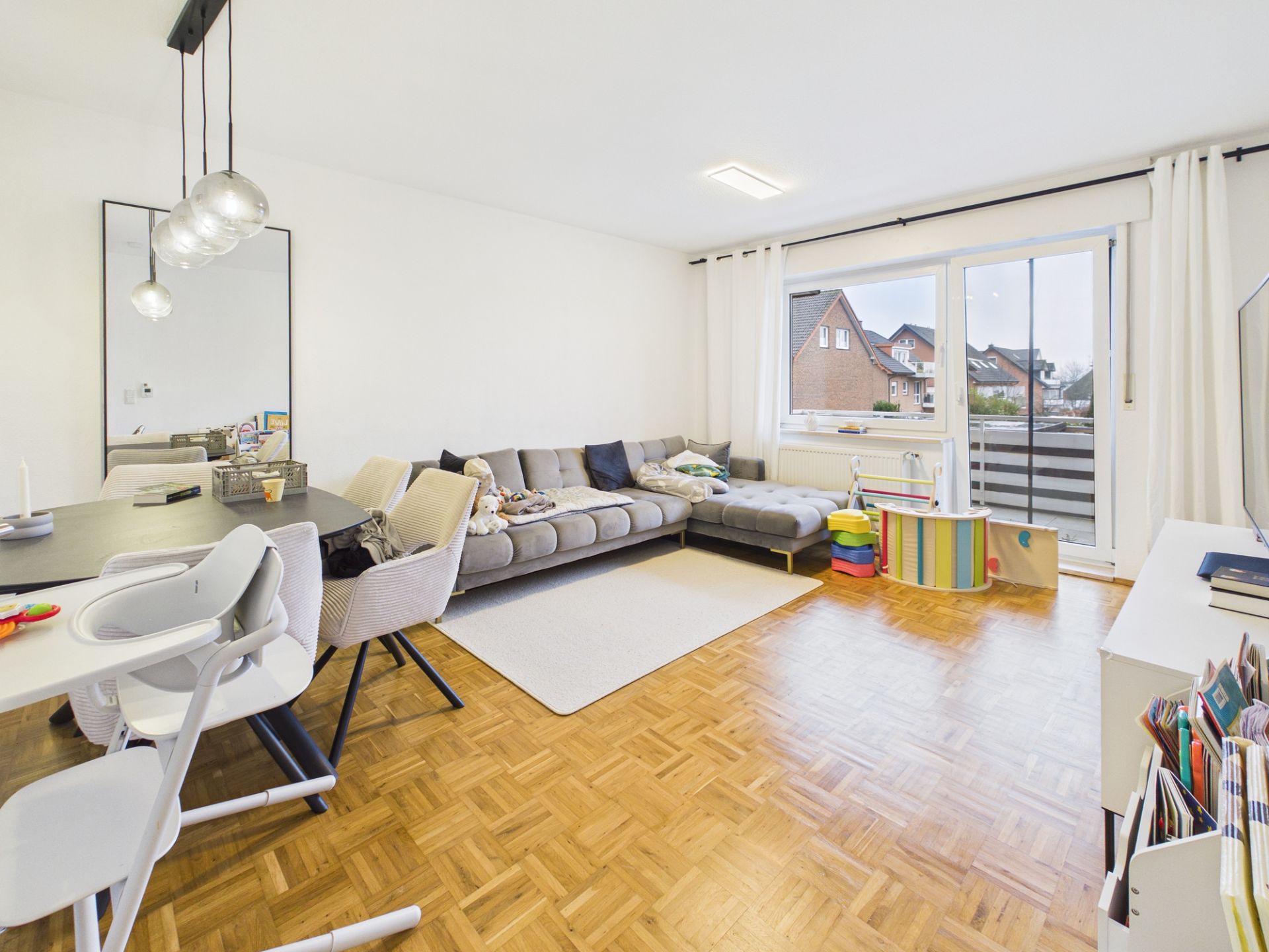 Helles Wohnzimmer mit grauem Sofa, Essbereich, Parkettboden und Zugang zum Balkon.