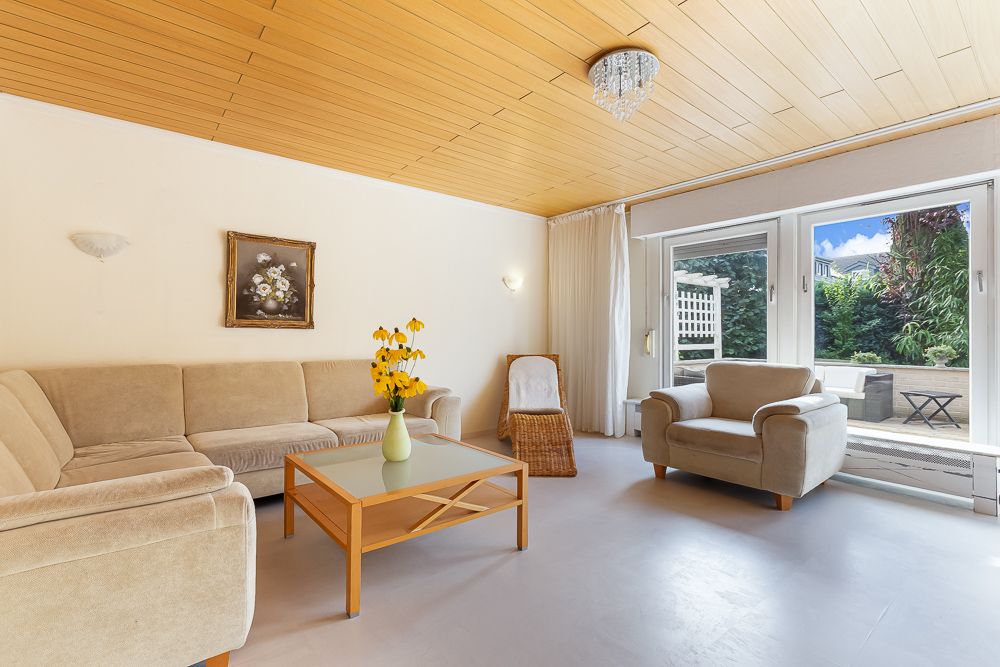 Helles Wohnzimmer mit Sofa, Sessel, Glastisch und Blick auf Terrasse mit Garten.