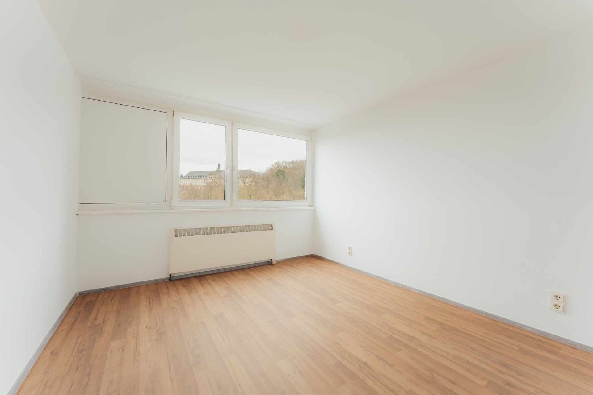 Leeres Zimmer mit weiß gestrichenen Wänden, Holzfußboden und großen Fenstern.