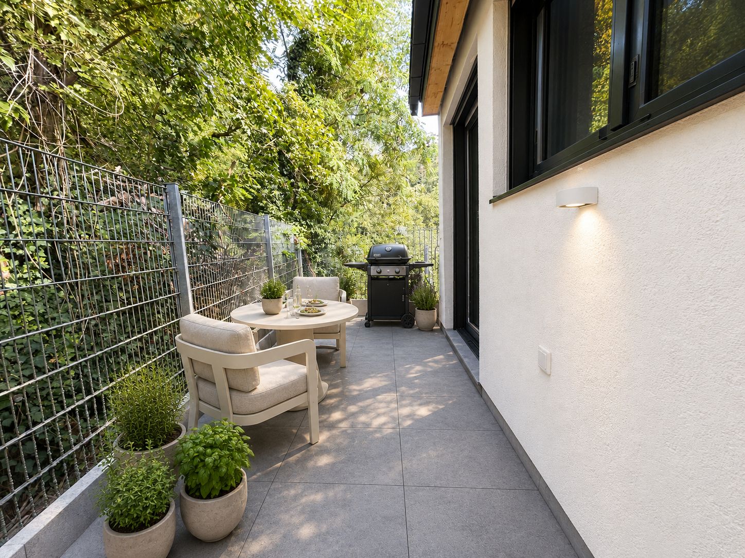 Kleine Terrasse mit zwei Sesseln, rundem Tisch, Pflanzen und Grill am Rand eines Hauses.