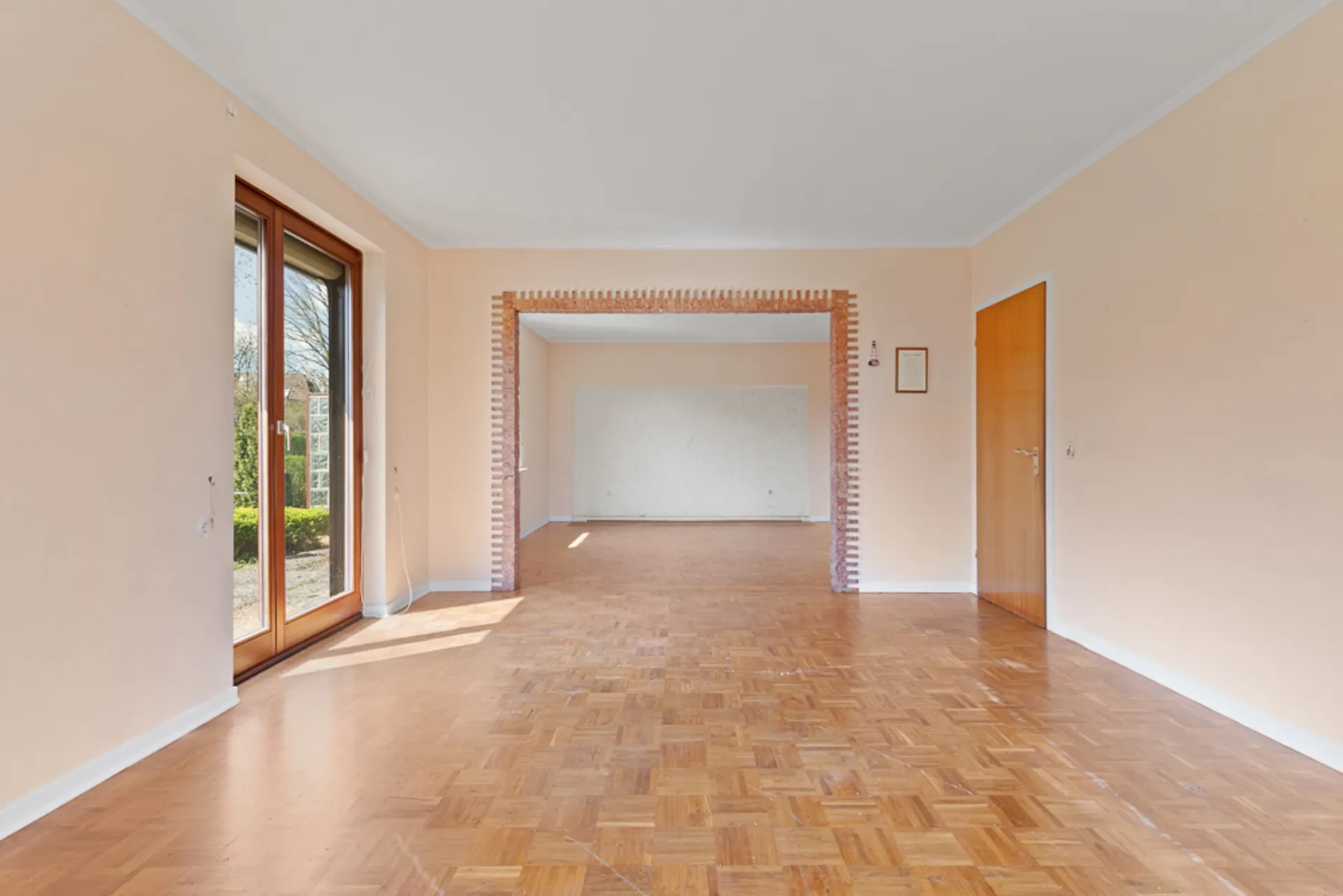Leeres Wohnzimmer mit Parkettboden, großen Fenstern und einer offenen Durchgangstür zum Nebenraum.