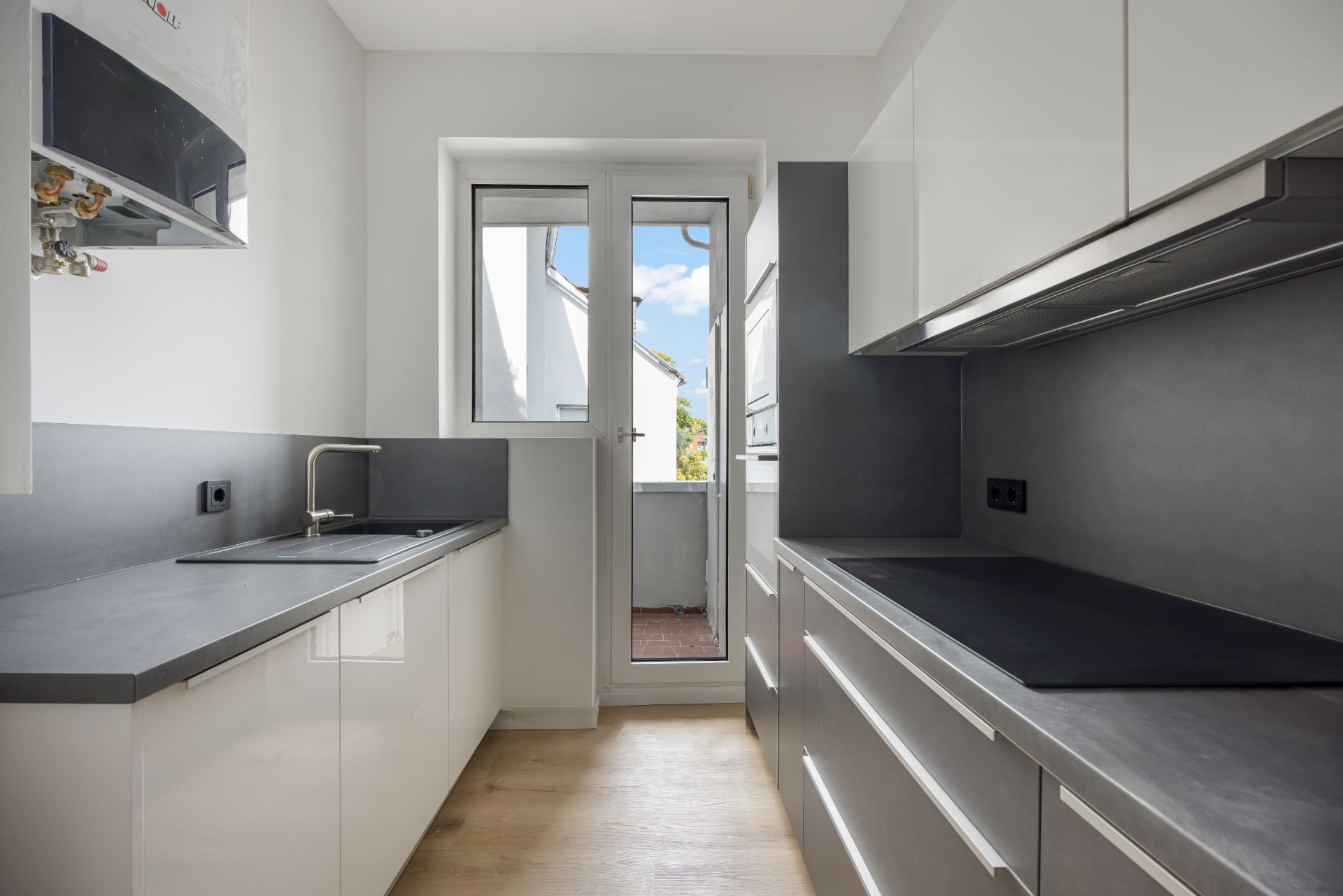 Moderne, schmale Küche mit weißen Schränken, grauen Arbeitsplatten und Zugang zu einem kleinen Balkon.