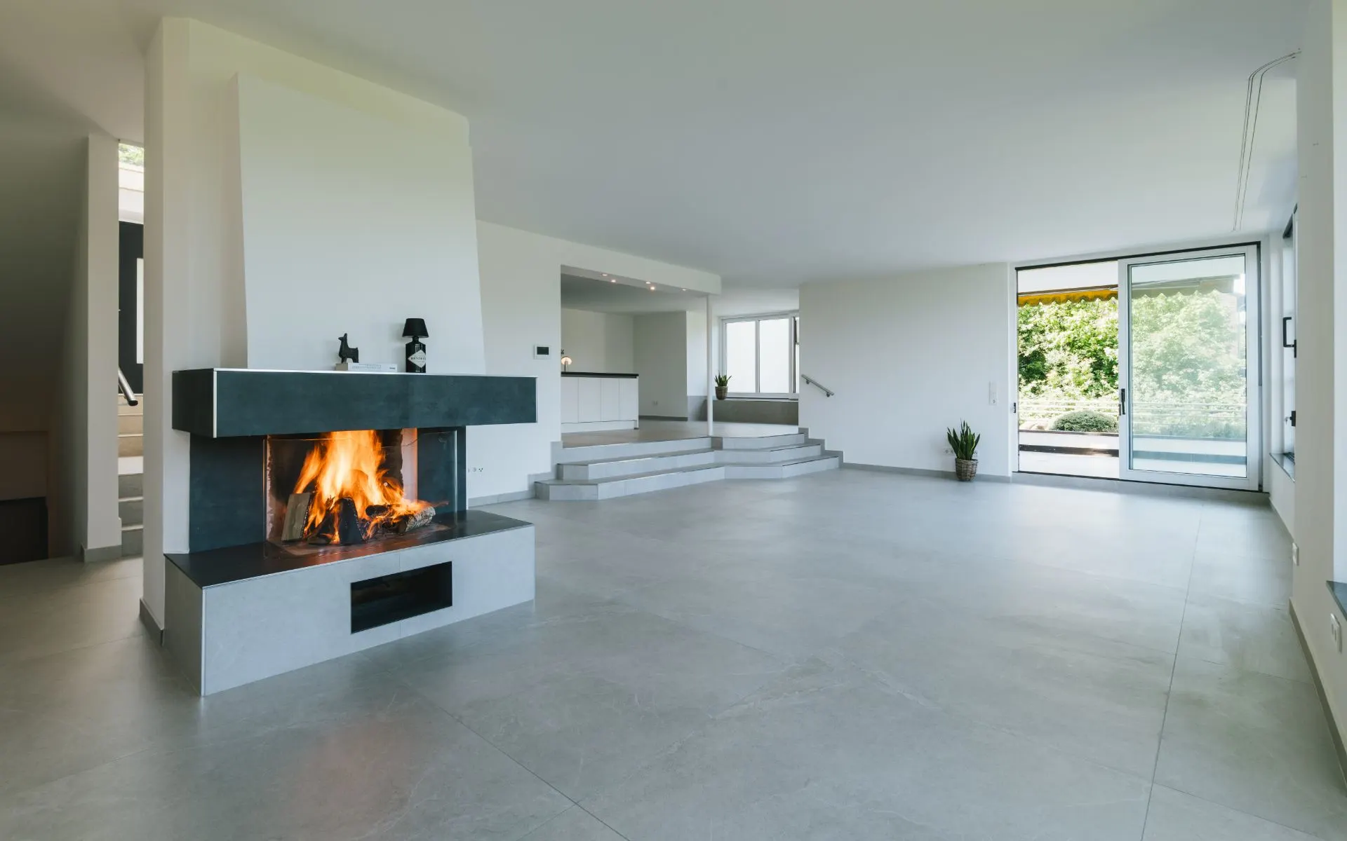 Großes, modernes Wohnzimmer mit Kamin, grauem Boden und Gartenzugang durch Glastüren.