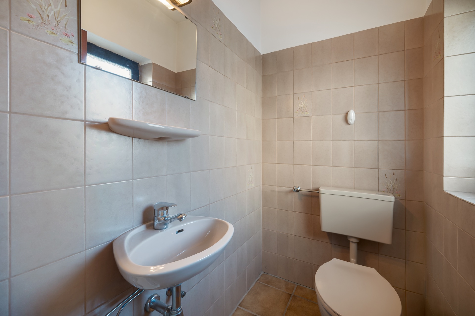 Kleines Badezimmer mit Waschbecken, Spiegel und Toilette, beigefarbene Fliesen an den Wänden.