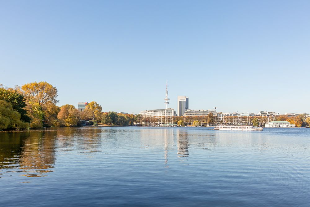 Großer ruhiger See mit Bäumen und Gebäuden im Hintergrund bei klarem Himmel.