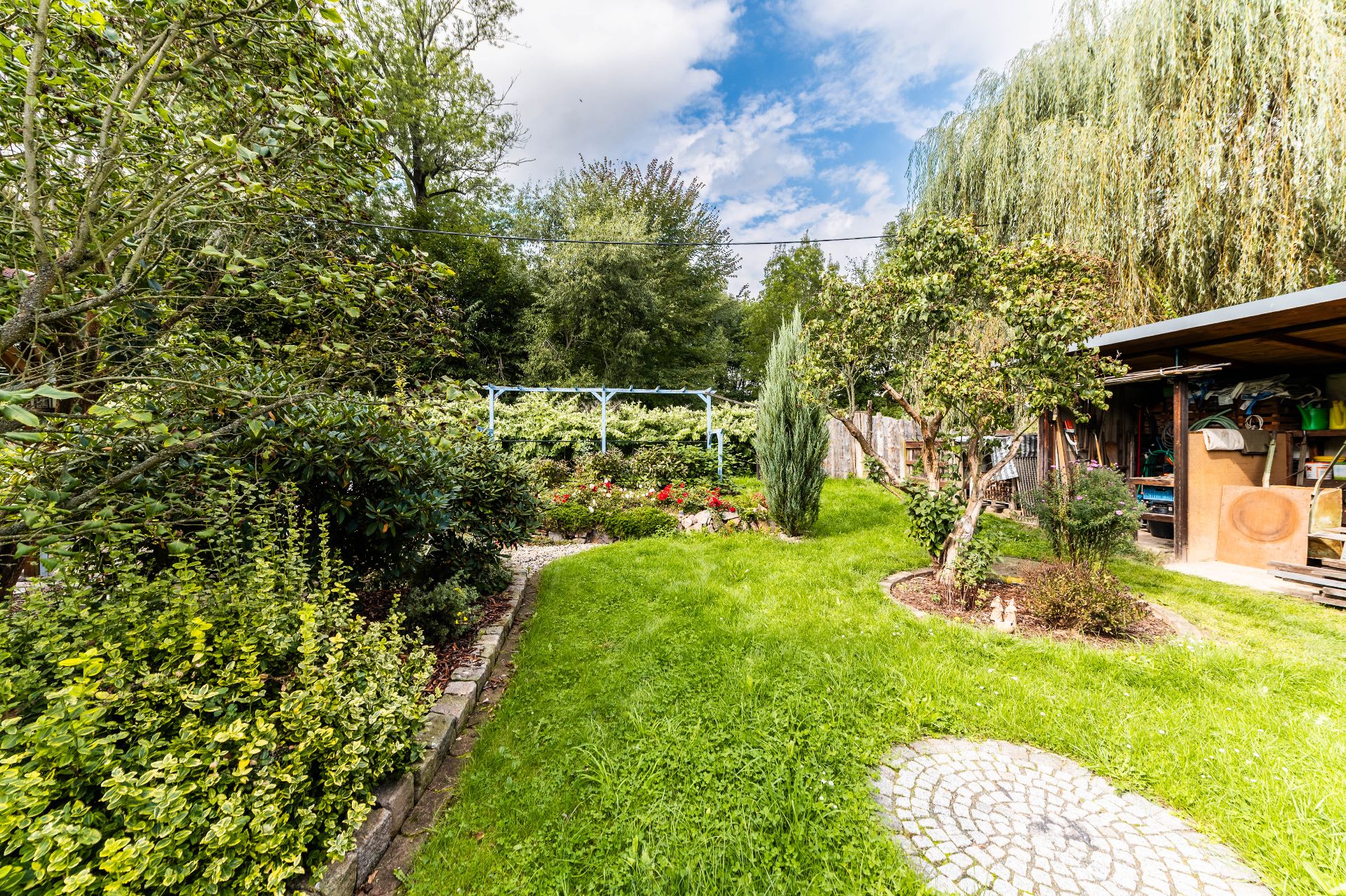 Grüner Garten mit Rasen, Wegen, Bäumen und einem überdachten Abstellbereich rechts.