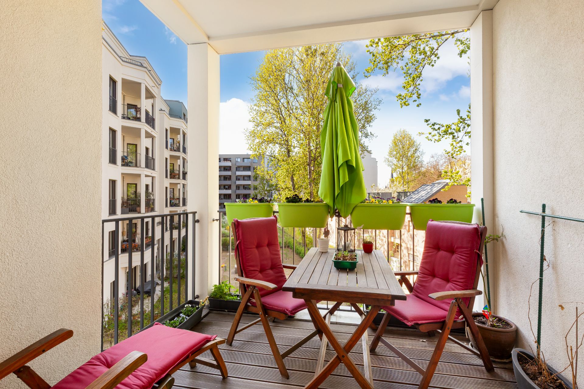 Balkon mit Holztisch, vier roten Stühlen, Pflanzen und grünem Sonnenschirm.