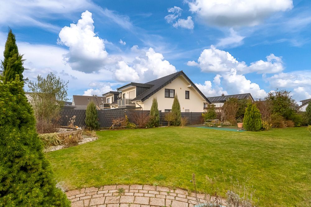 Großer Garten mit Rasen, Bäumen und Blick auf ein Einfamilienhaus unter blauem Himmel.