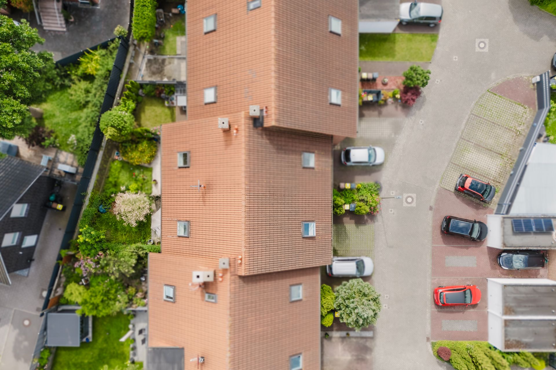 Luftaufnahme eines Wohnhauses mit rotbraunem Dach, umgeben von Gärten und parkenden Autos.