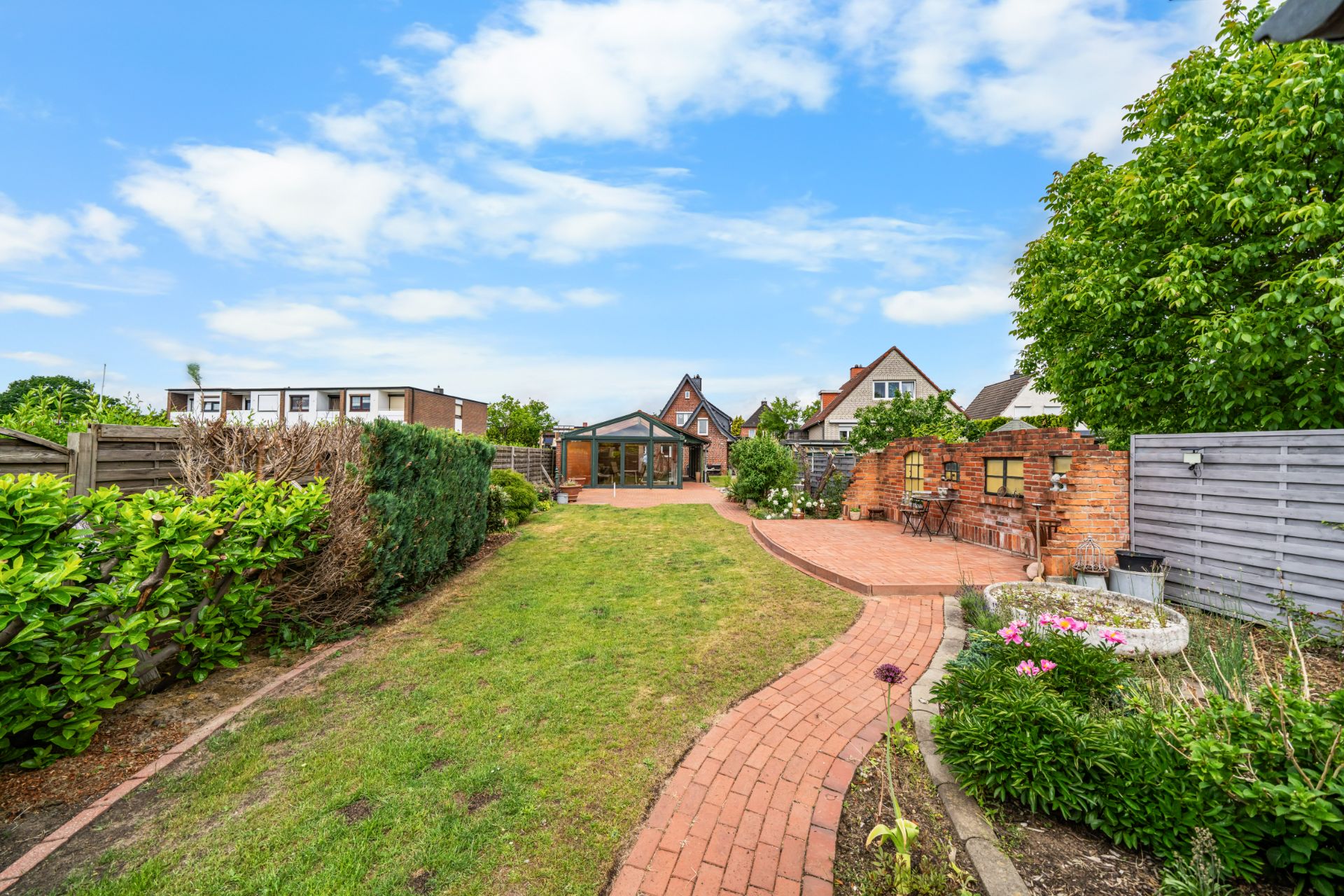 Garten mit Rasenfläche, Blumenbeeten, Weg aus roten Steinen und Terrasse im Hintergrund.