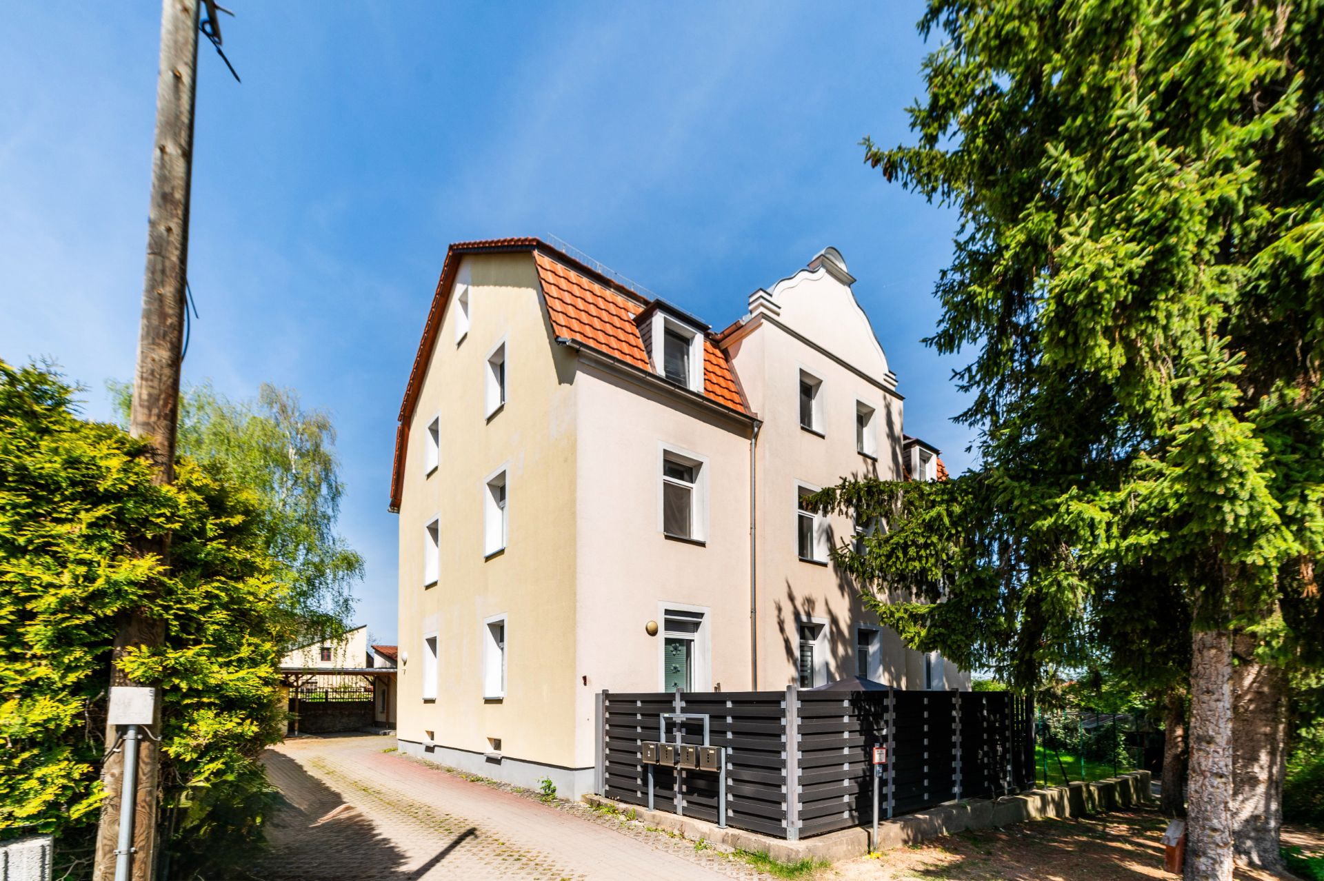 Mehrstöckiges Wohnhaus mit beigem Anstrich und rotem Dach, umgeben von Bäumen.
