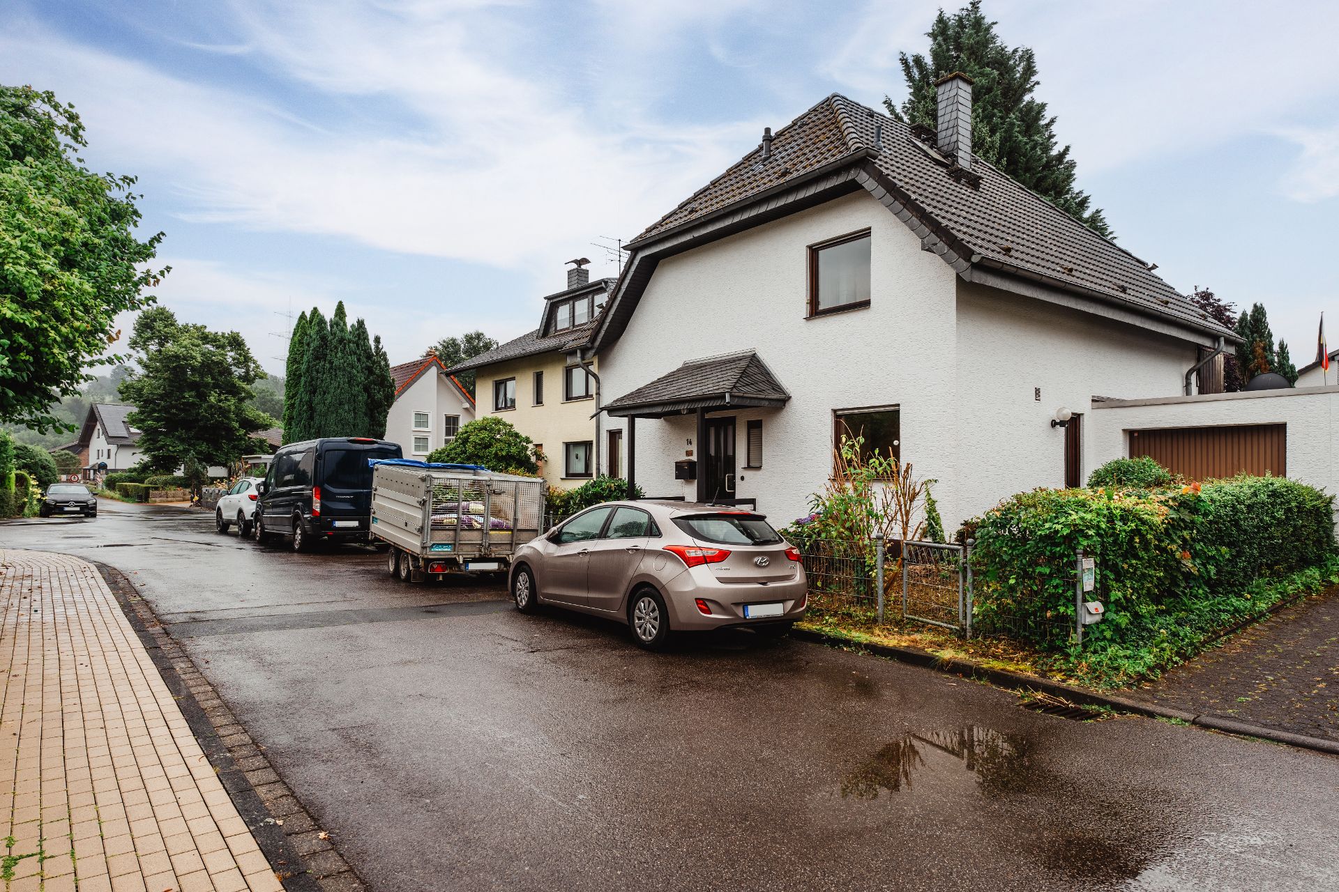 Wohnstraße mit Einfamilienhäusern, parkenden Autos und Anhänger bei nassem Wetter.