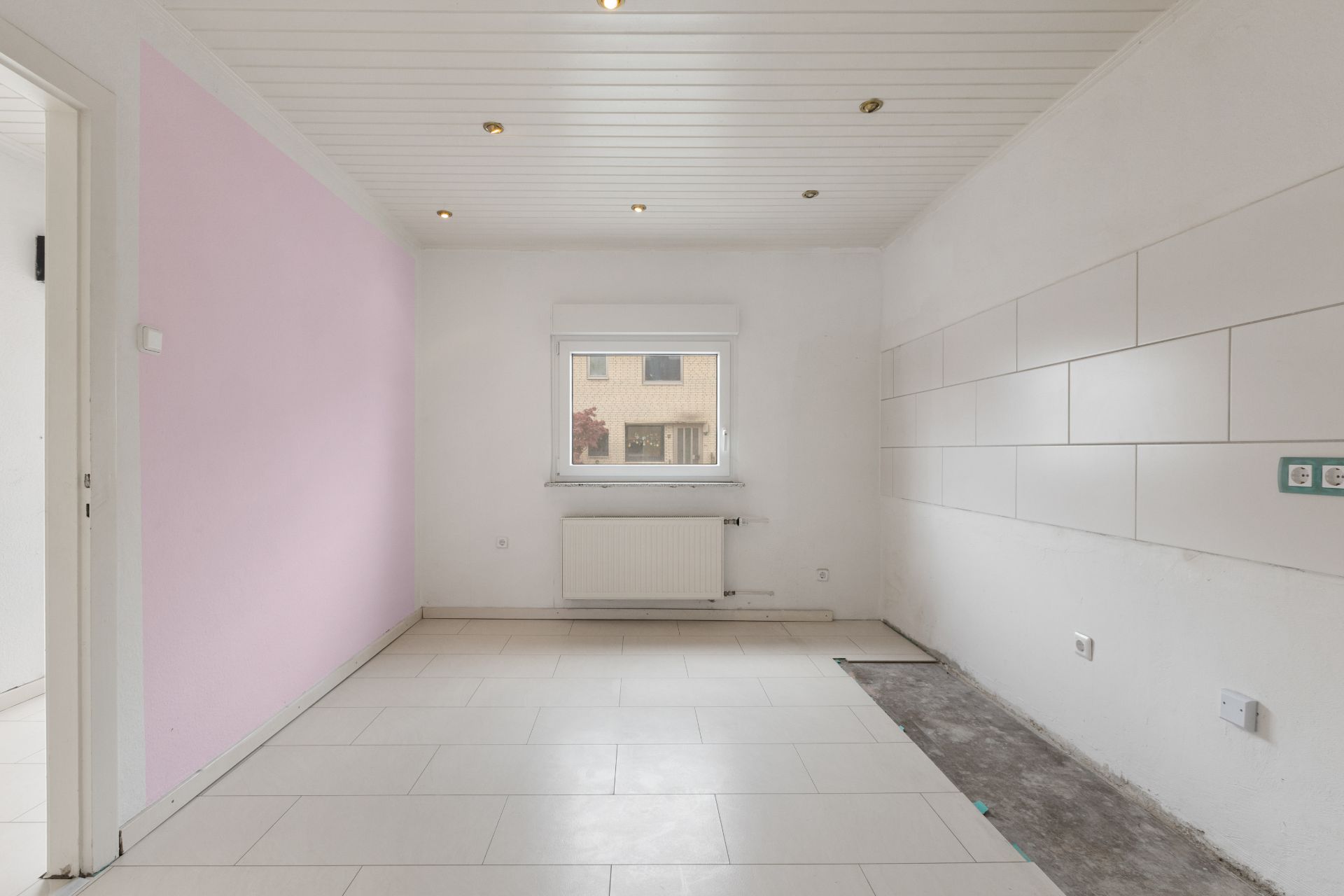 Leerer Raum mit weißen Fliesen, rosa Wand und Fenster, Steckdosen an der rechten Wand.