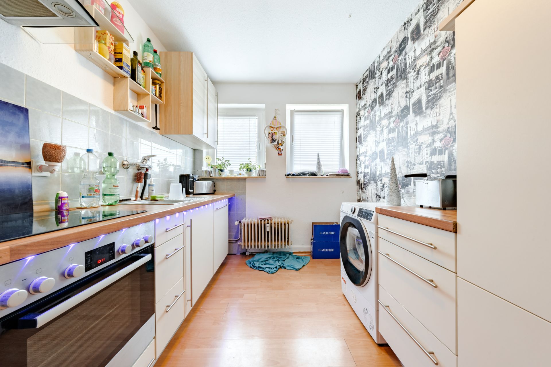 Helle Küche mit Herd, Waschmaschine, Regalen, zwei Fenstern und gemusterter Wand.