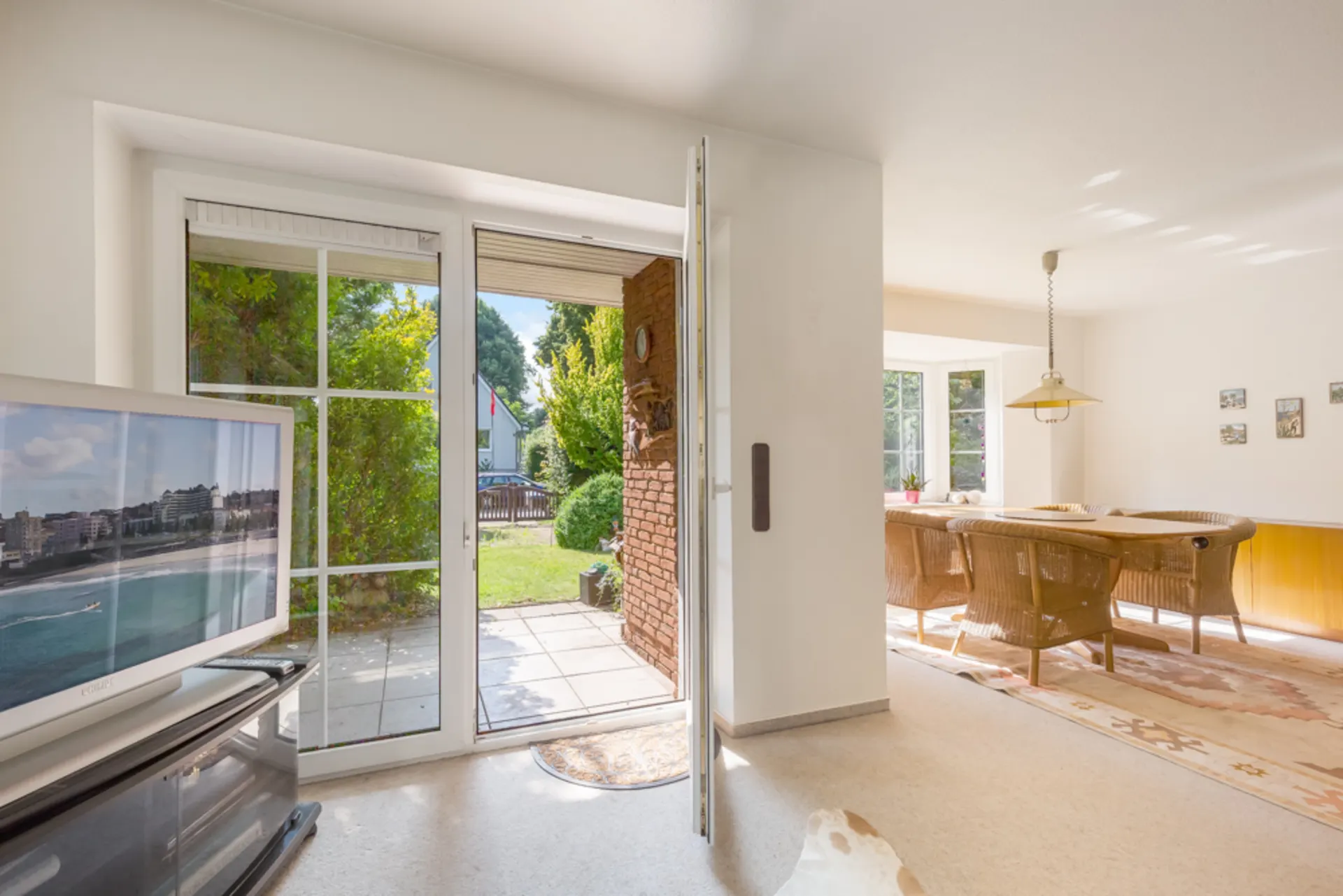 Helles Wohnzimmer mit Fernseher, Esstisch und geöffneter Tür zur Terrasse und Garten.
