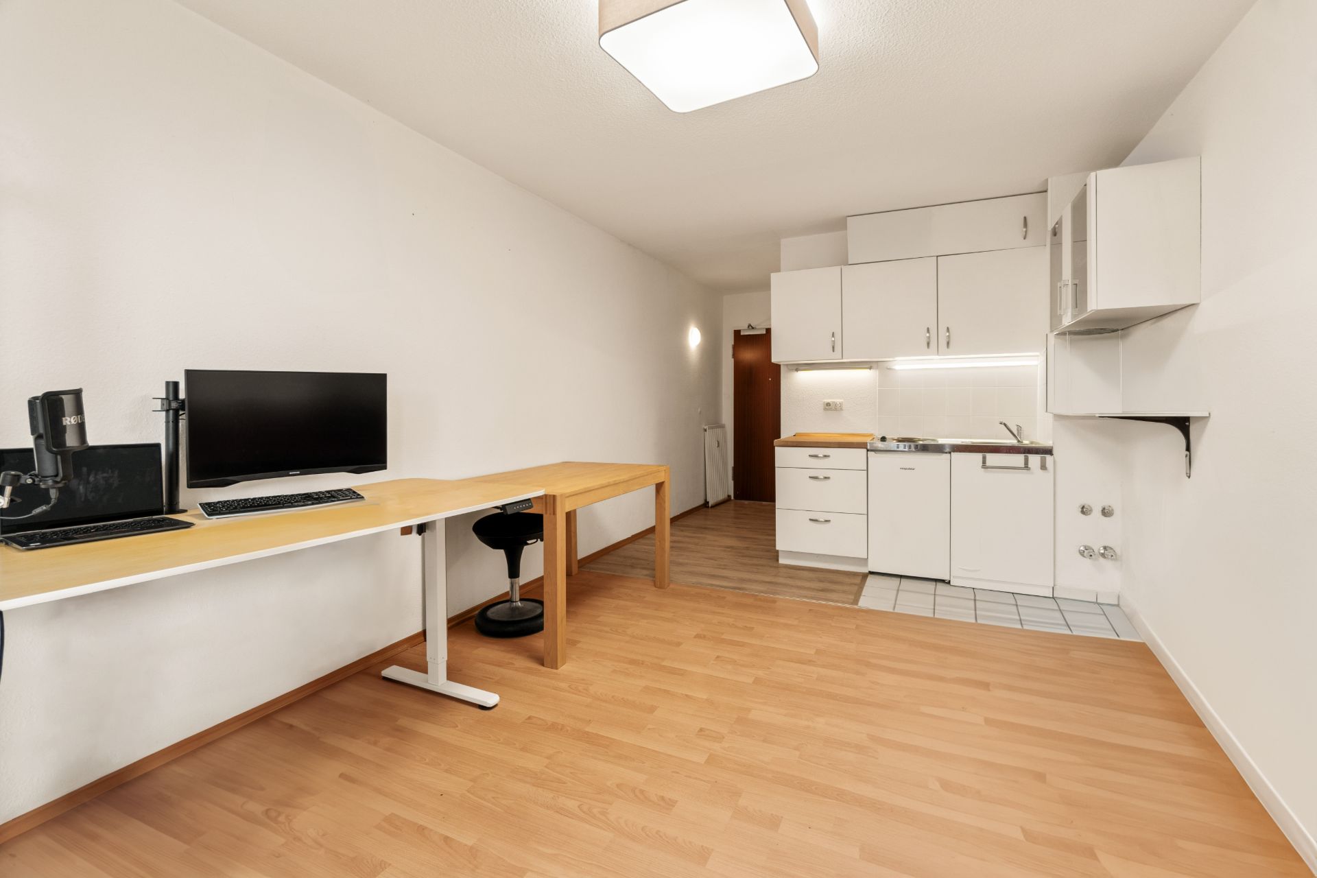 Helles Zimmer mit Schreibtisch, Computer und kleiner Küchenzeile auf Laminatboden.