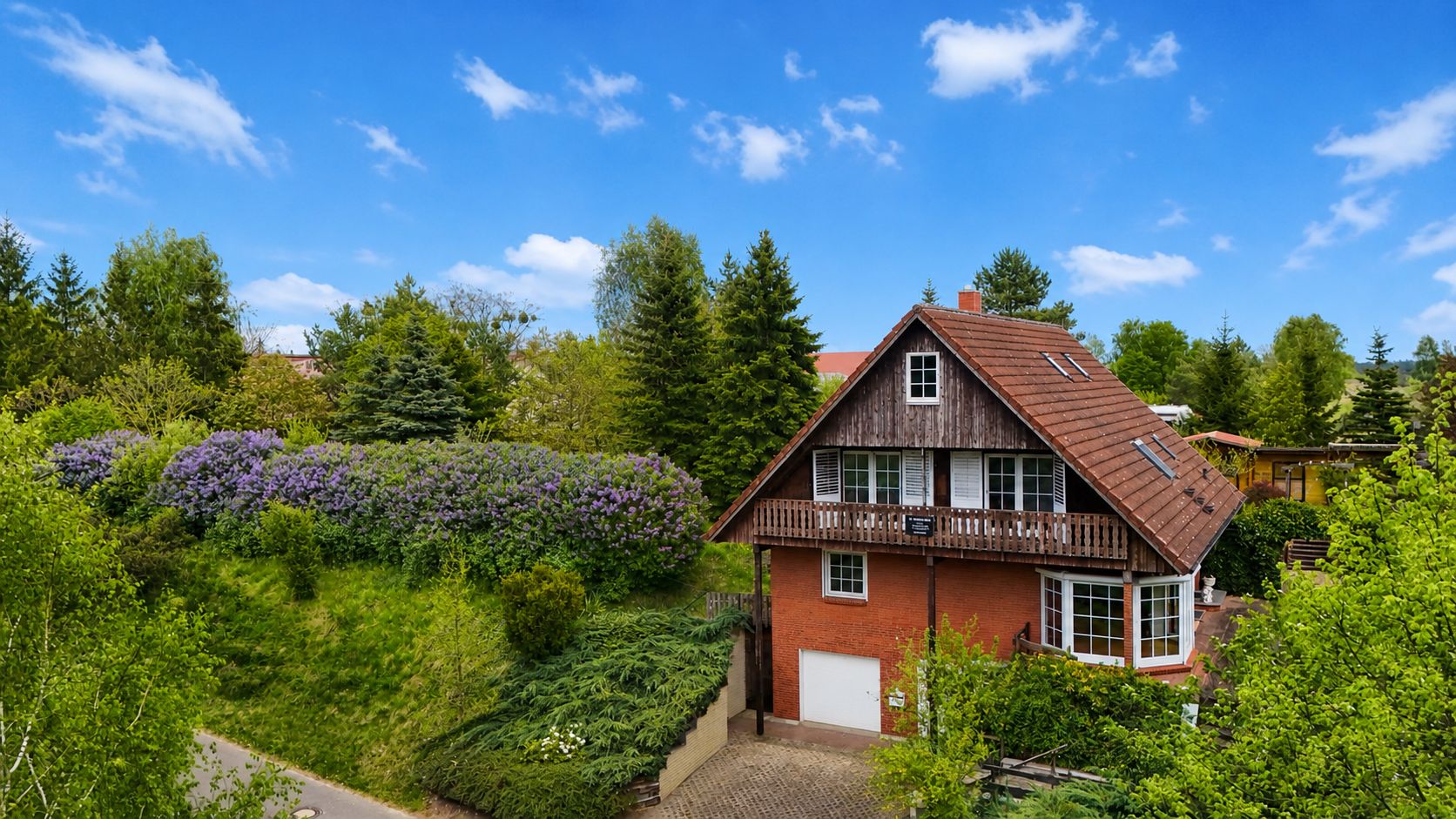 Ein rotes Einfamilienhaus mit Balkon, umgeben von Bäumen und viel Grün bei blauem Himmel.
