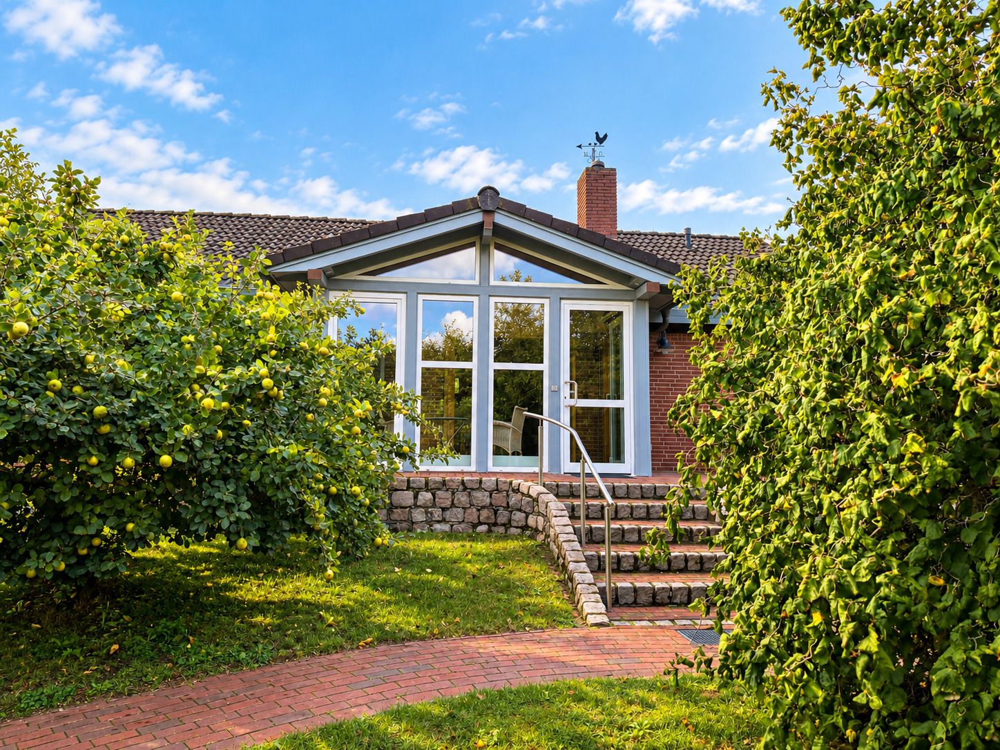 Eingang eines Hauses mit Glasfront, Steinmauer und Treppe, umgeben von Bäumen im Garten.