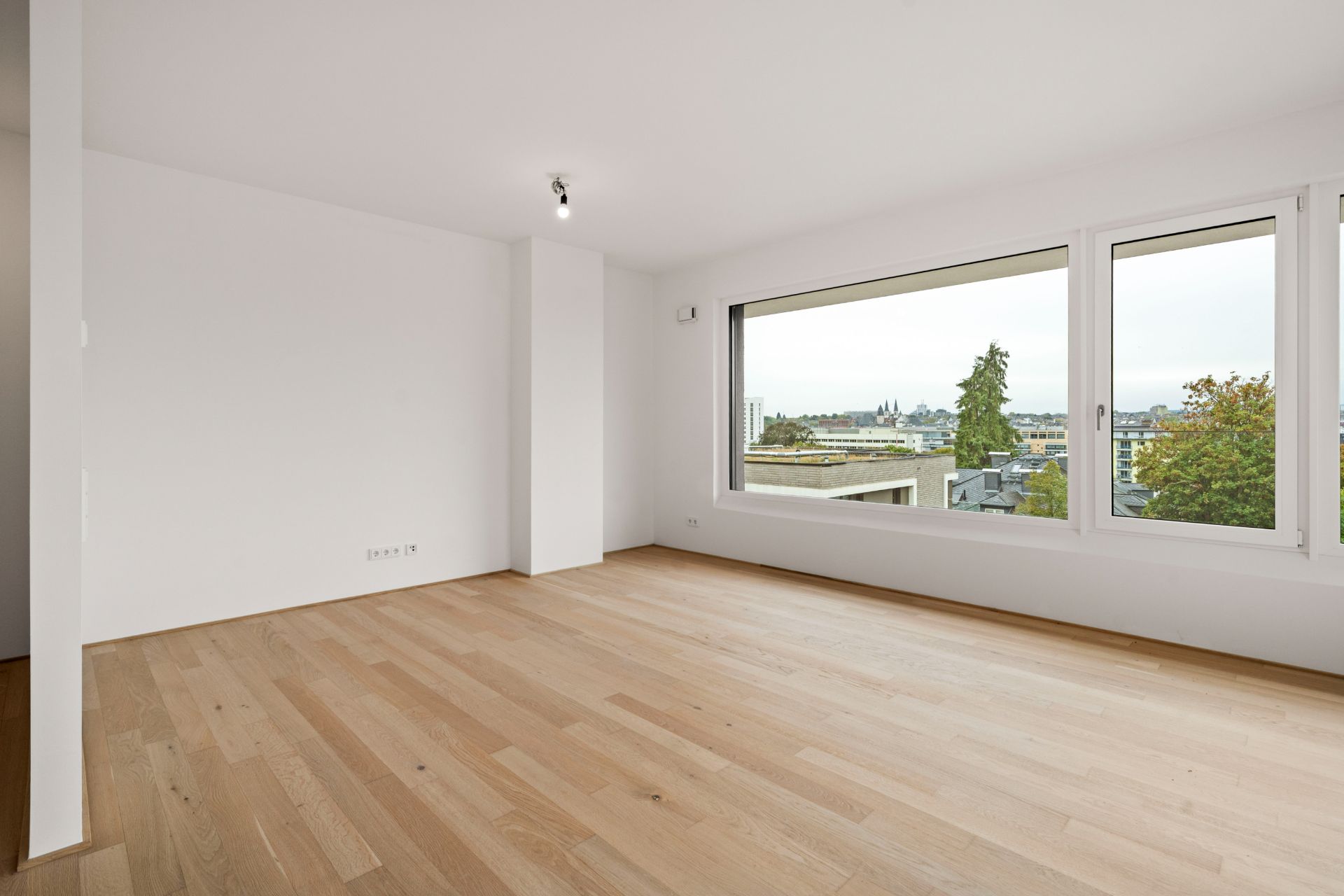 Helles, leeres Zimmer mit Holzfußboden und großem Fenster mit Blick nach draußen.