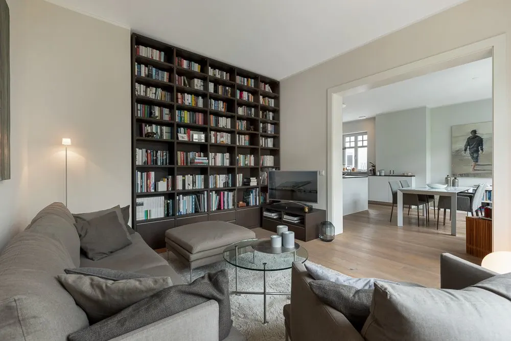 Wohnzimmer mit grauem Sofa, Bücherregal, Fernseher und Blick in ein Esszimmer.