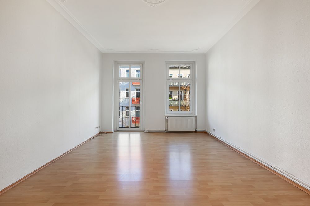 Helles, leeres Zimmer mit Parkettboden, zwei Fenstern und einer Glastür zu einem kleinen Balkon.