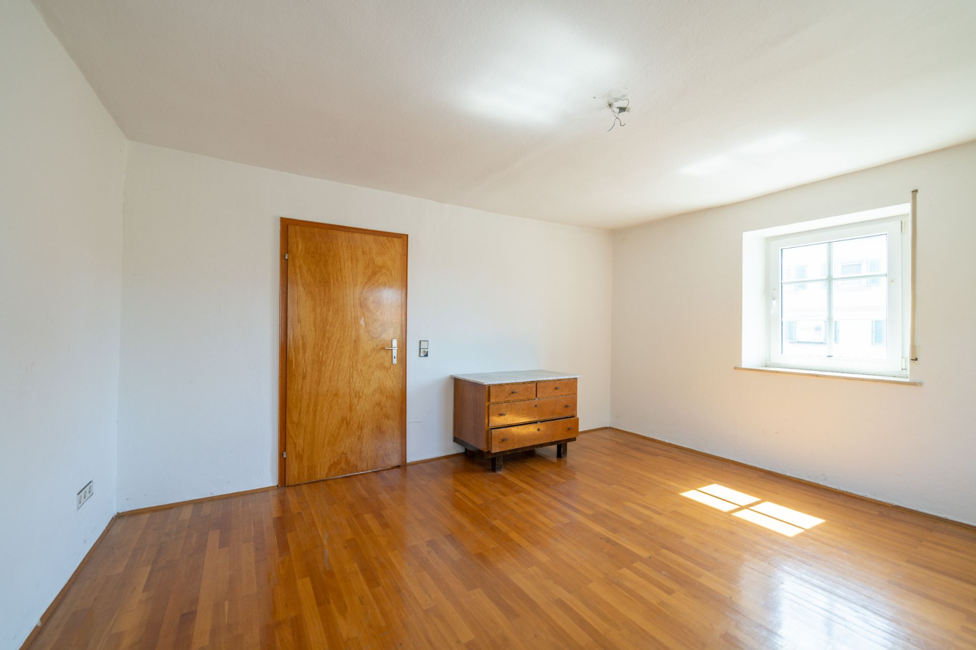 Leerer Raum mit Holzboden, Holztür, Kommode und Fenster mit Tageslicht.