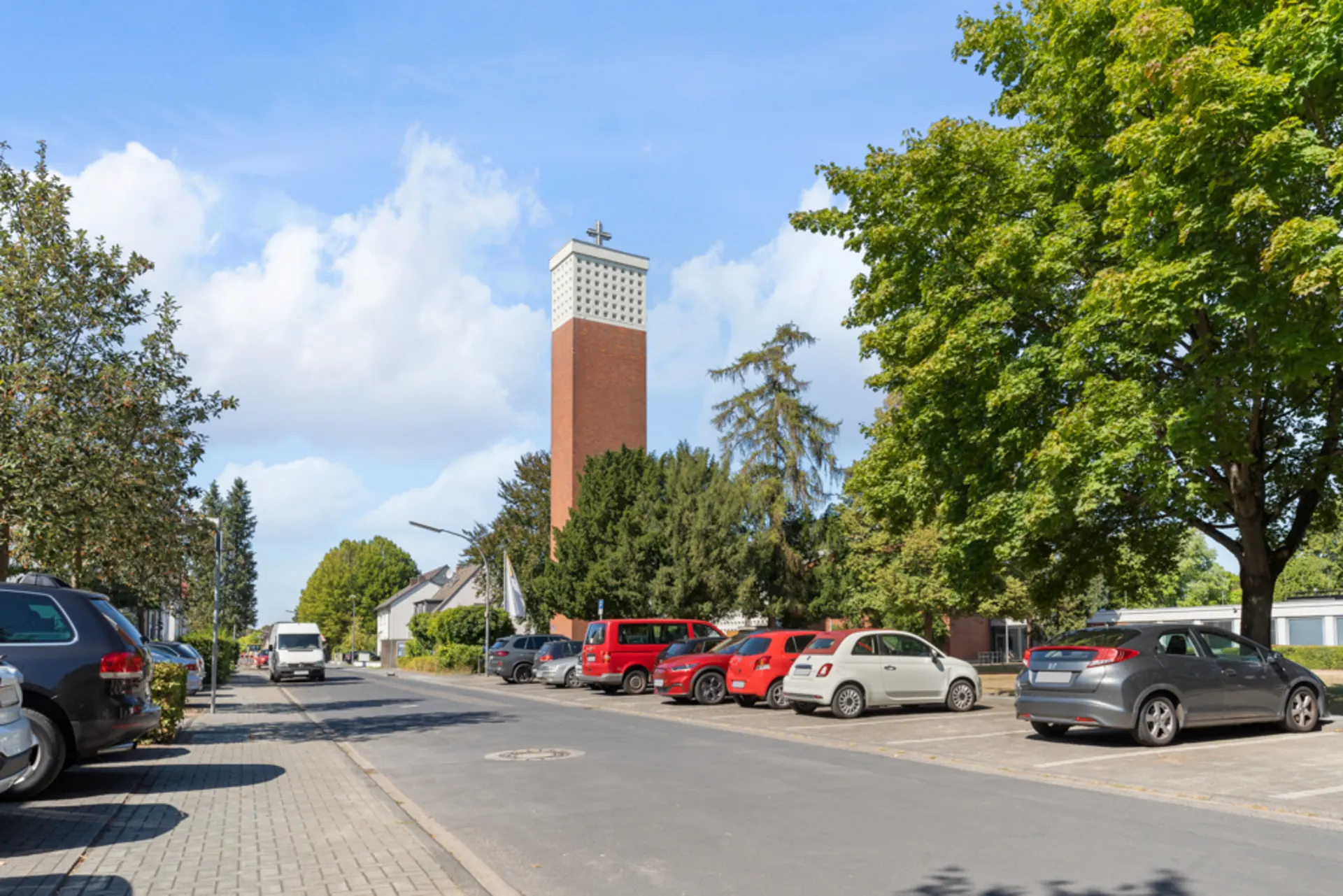 Straße mit parkenden Autos und Bäumen, im Hintergrund ein hoher Kirchturm.