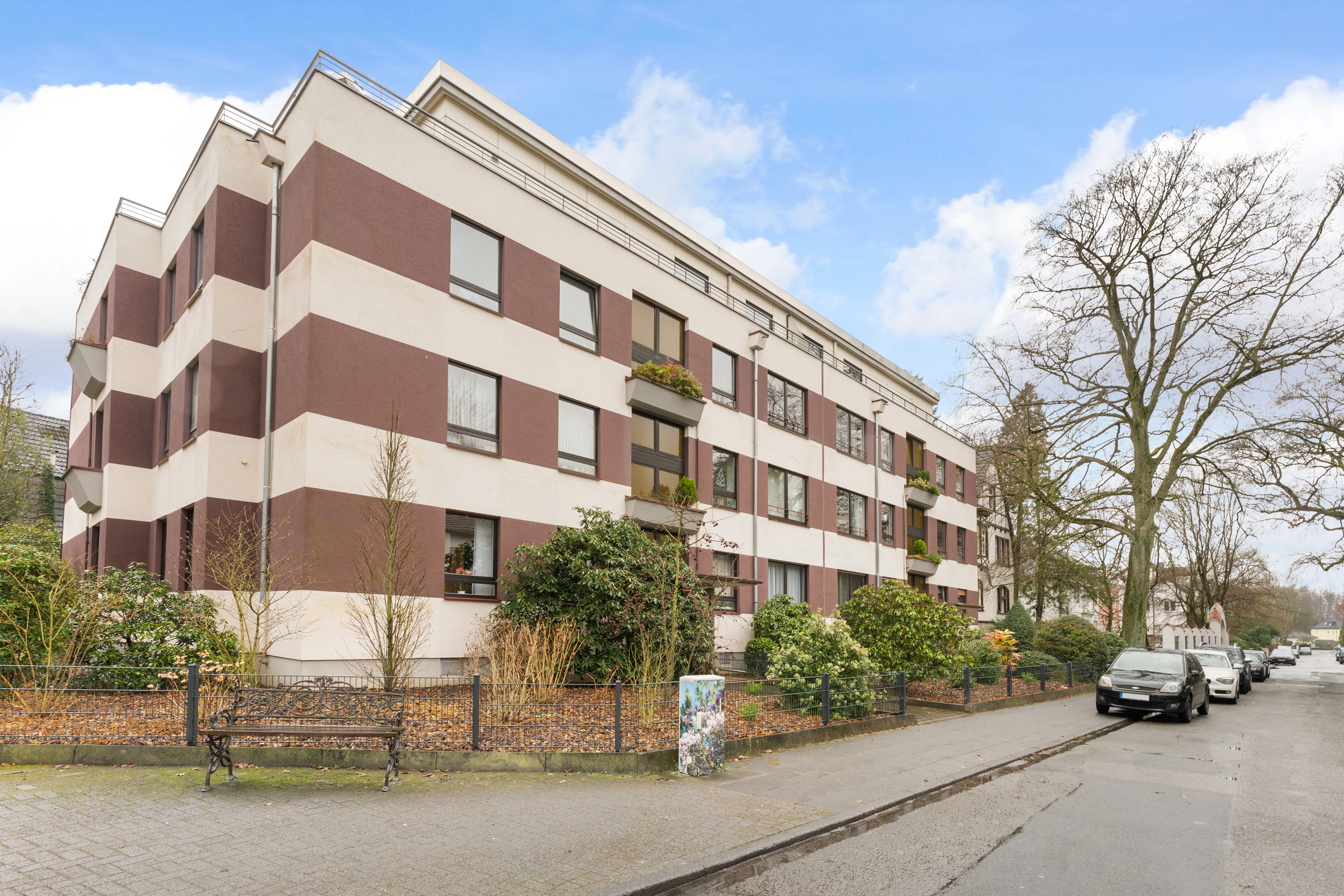 Mehrstöckiges Wohnhaus mit braunen und weißen Fassadenstreifen, Vorgarten und geparkten Autos an der Straße.