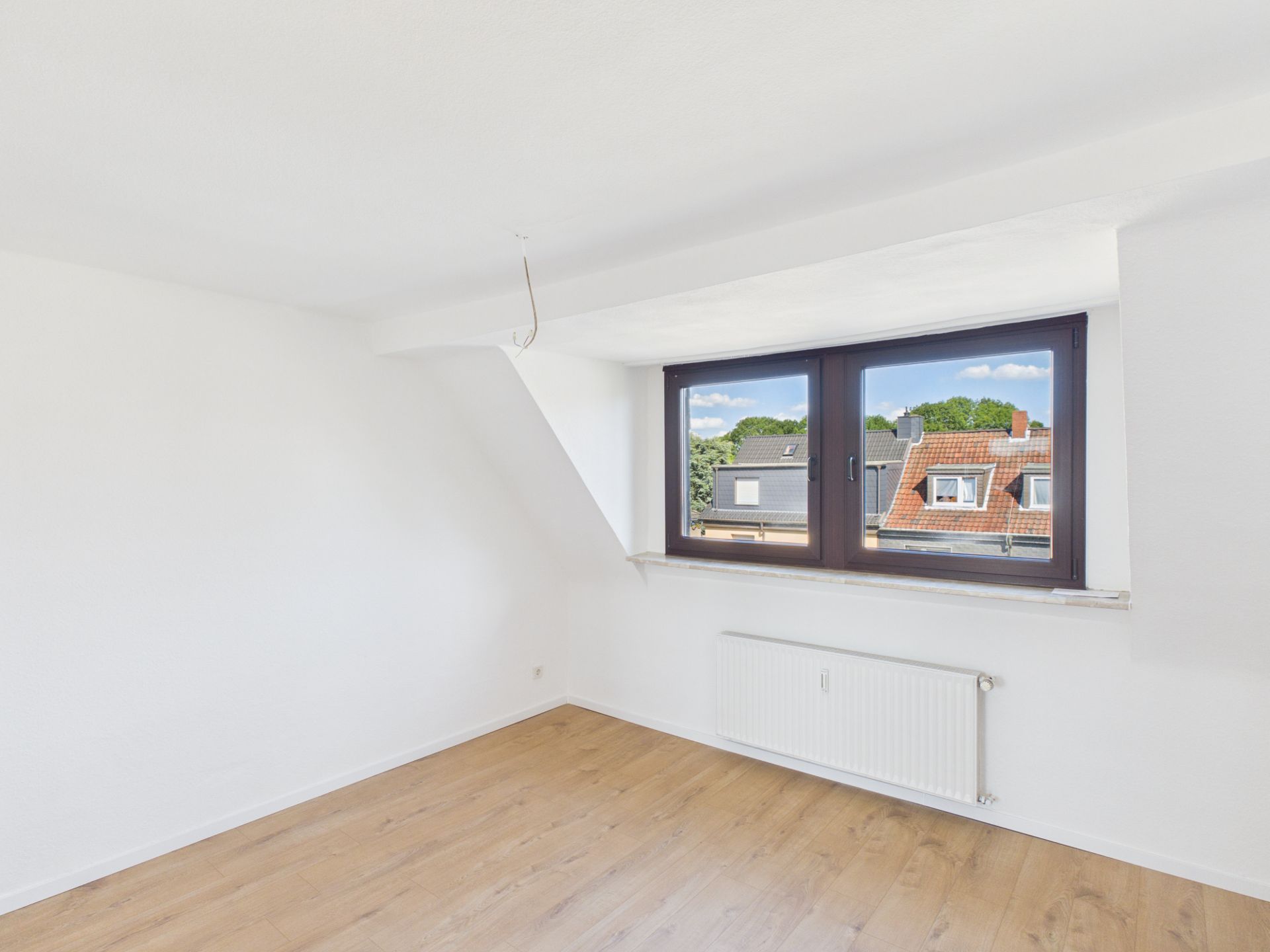 Helles Zimmer mit weißer Wand, Holzfußboden und großem Fenster mit Blick auf Nachbarhäuser.