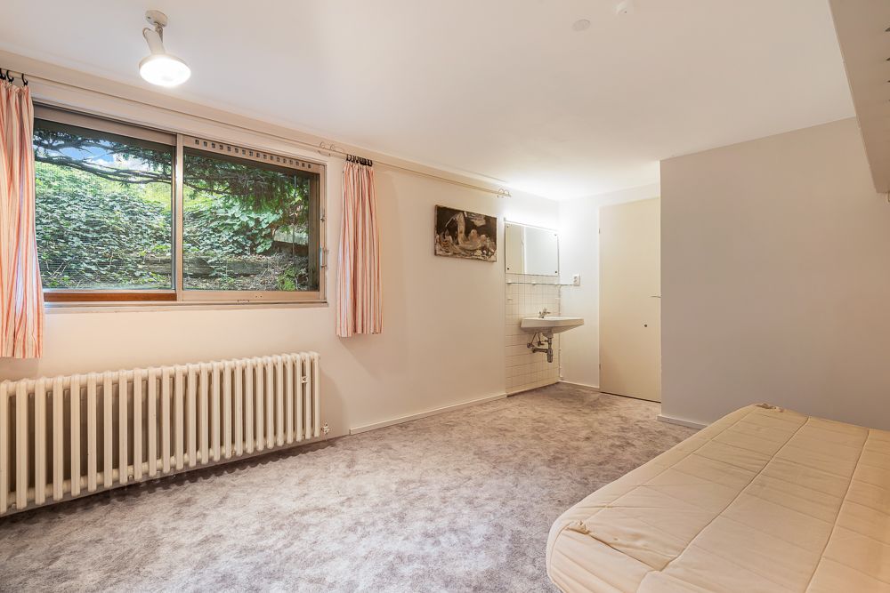 Helles Zimmer mit großem Fenster, Heizung, Teppichboden, Waschbecken und Bett.