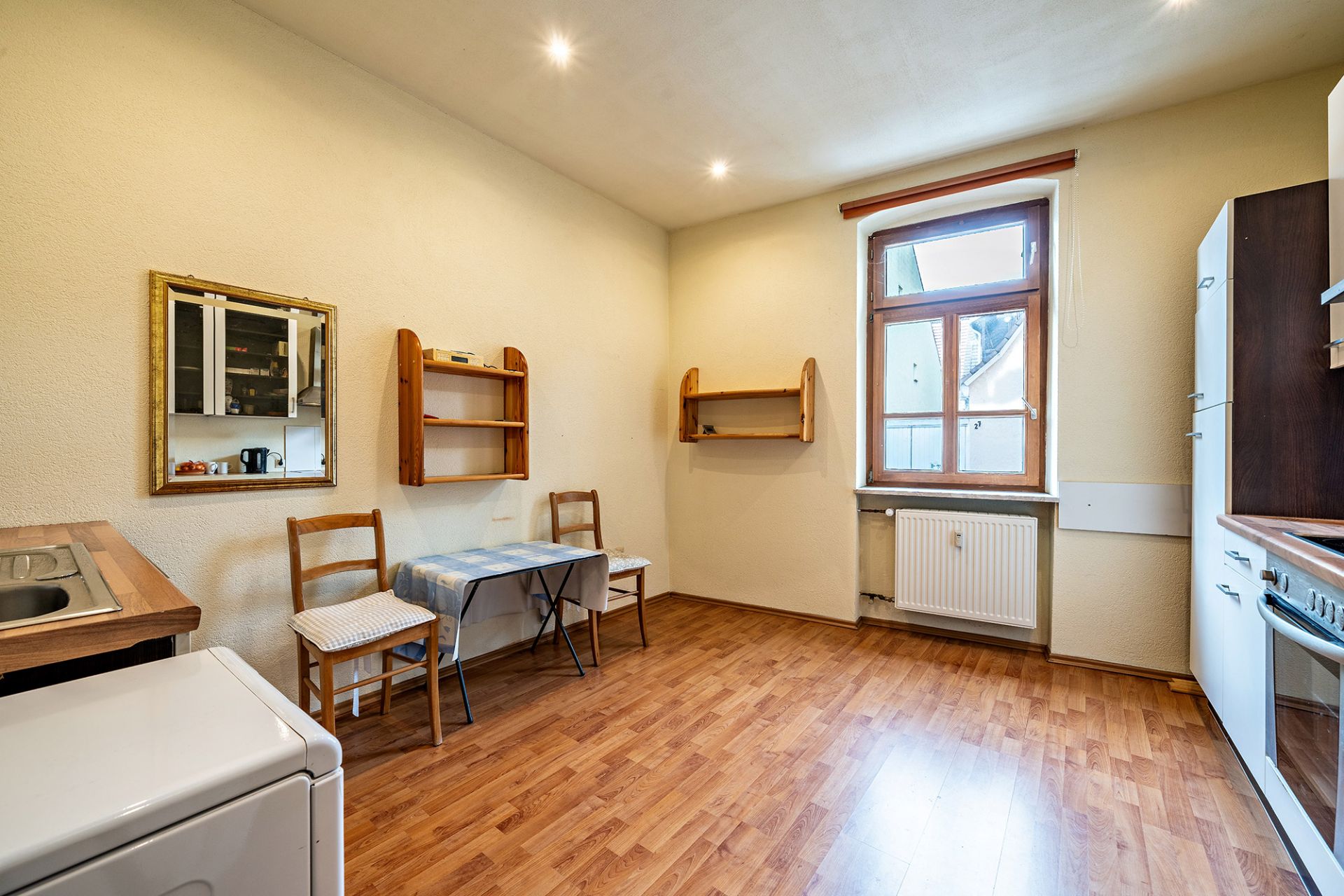 Helle Küche mit Holzboden, kleinem Esstisch, zwei Stühlen, Küchenschränken und Fenster.
